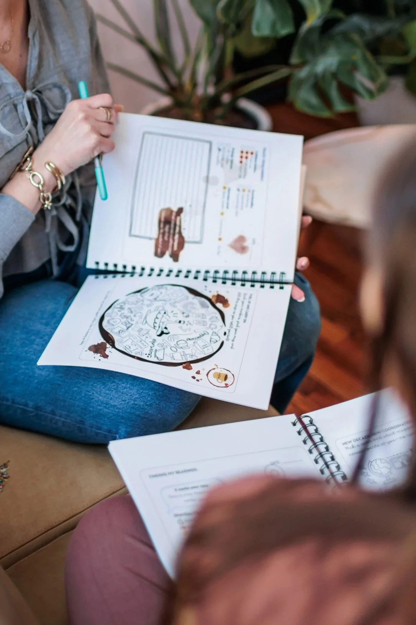 A person sitting on a couch or chair, holding and reading an open illustrated workbook with a coloring page and line drawings. The person is wearing jewelry, including bracelets and rings, and is dressed in a gray top and pink pants. A large plant with green leaves is in the background.