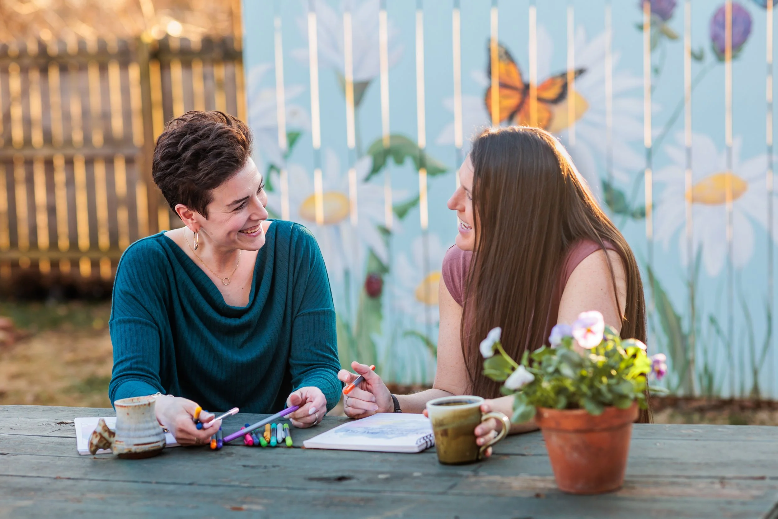 Two women sitting at a wooden table outdoors, smiling and talking to each other. The table has markers, a notebook, and coffee cups. In the background, there is a fence with large painted flowers and a butterfly.