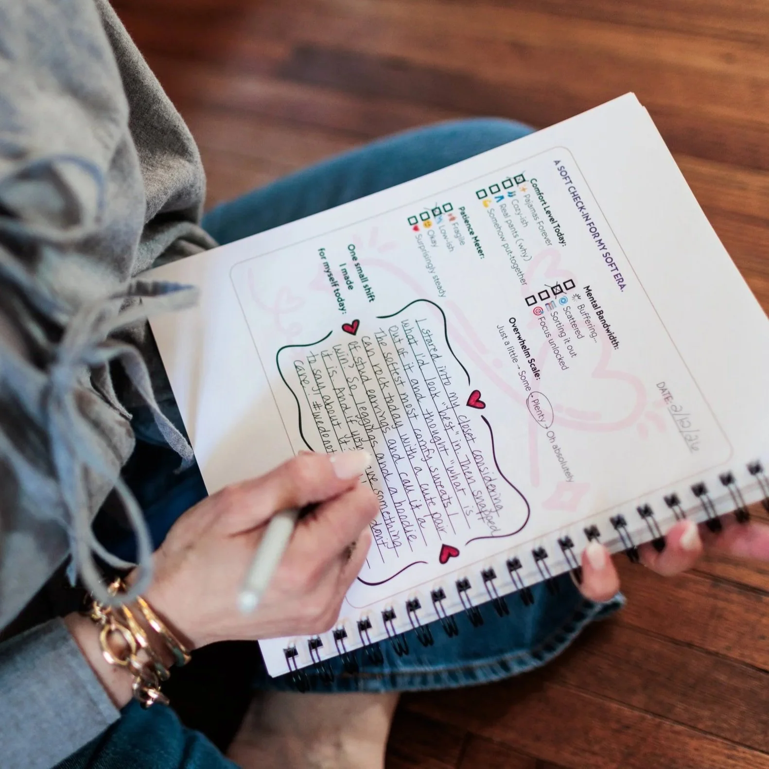 Person holding a spiral-bound notebook with handwritten notes, decorated with doodles and hearts, sitting on a wooden surface.