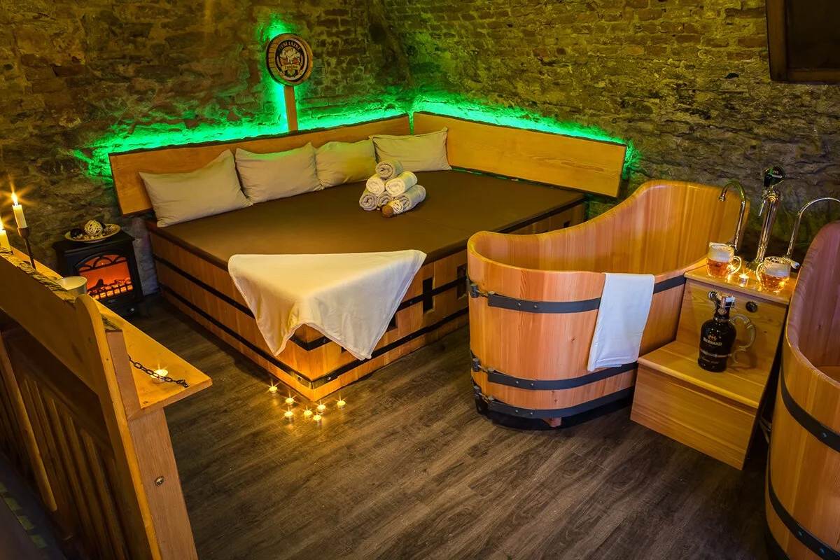 A cozy room with stone walls and wooden furniture, including a bed with rolled towels, a bathtub with a towel hanging over the side, and small candles illuminating the space.