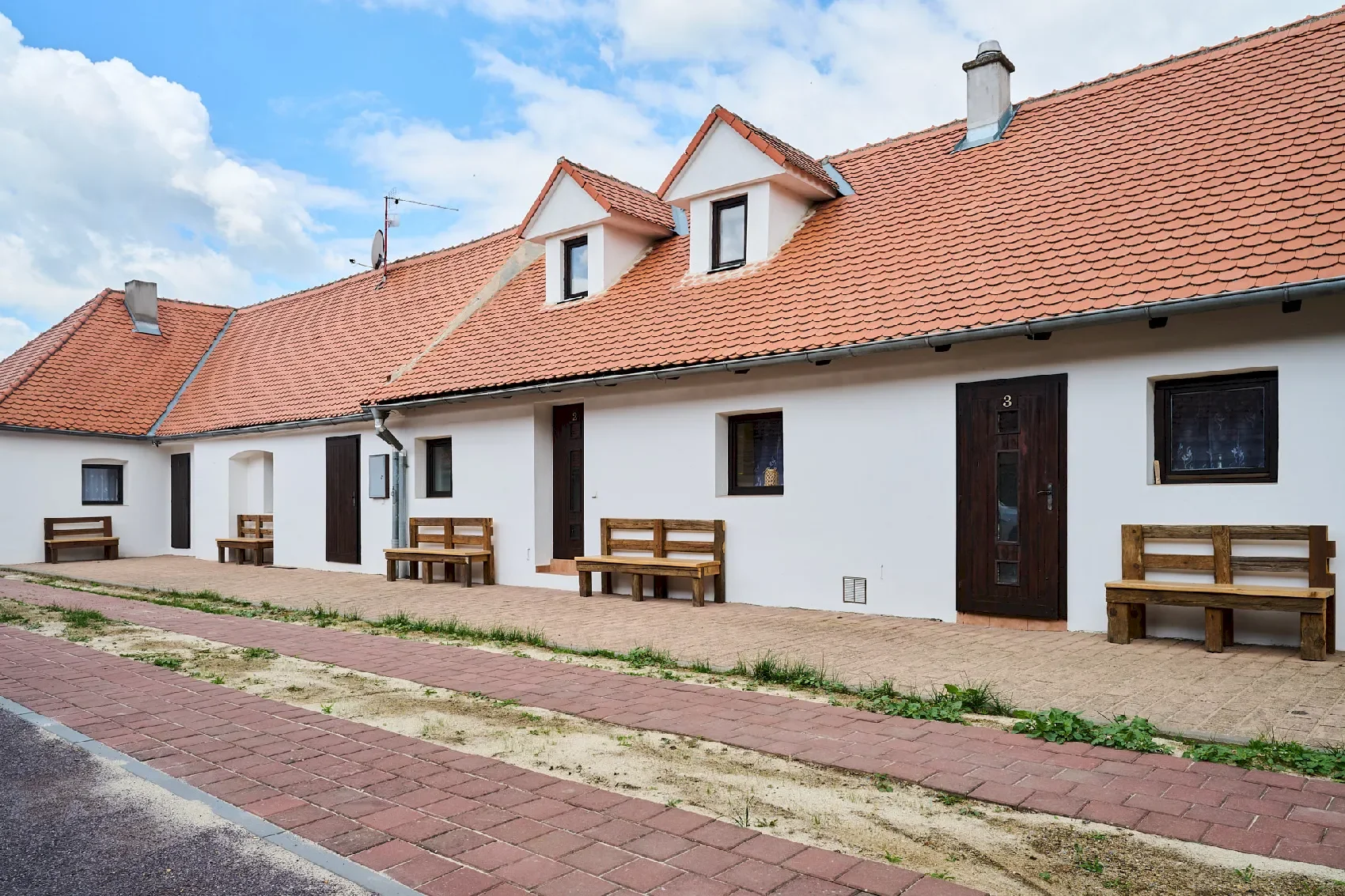 A residential building with a white exterior, black window frames, and a red-tiled roof. Multiple wooden benches are placed along the front of the building, and there's a paved walkway in front of the benches. The sky is partly cloudy with some clouds and blue sky visible.