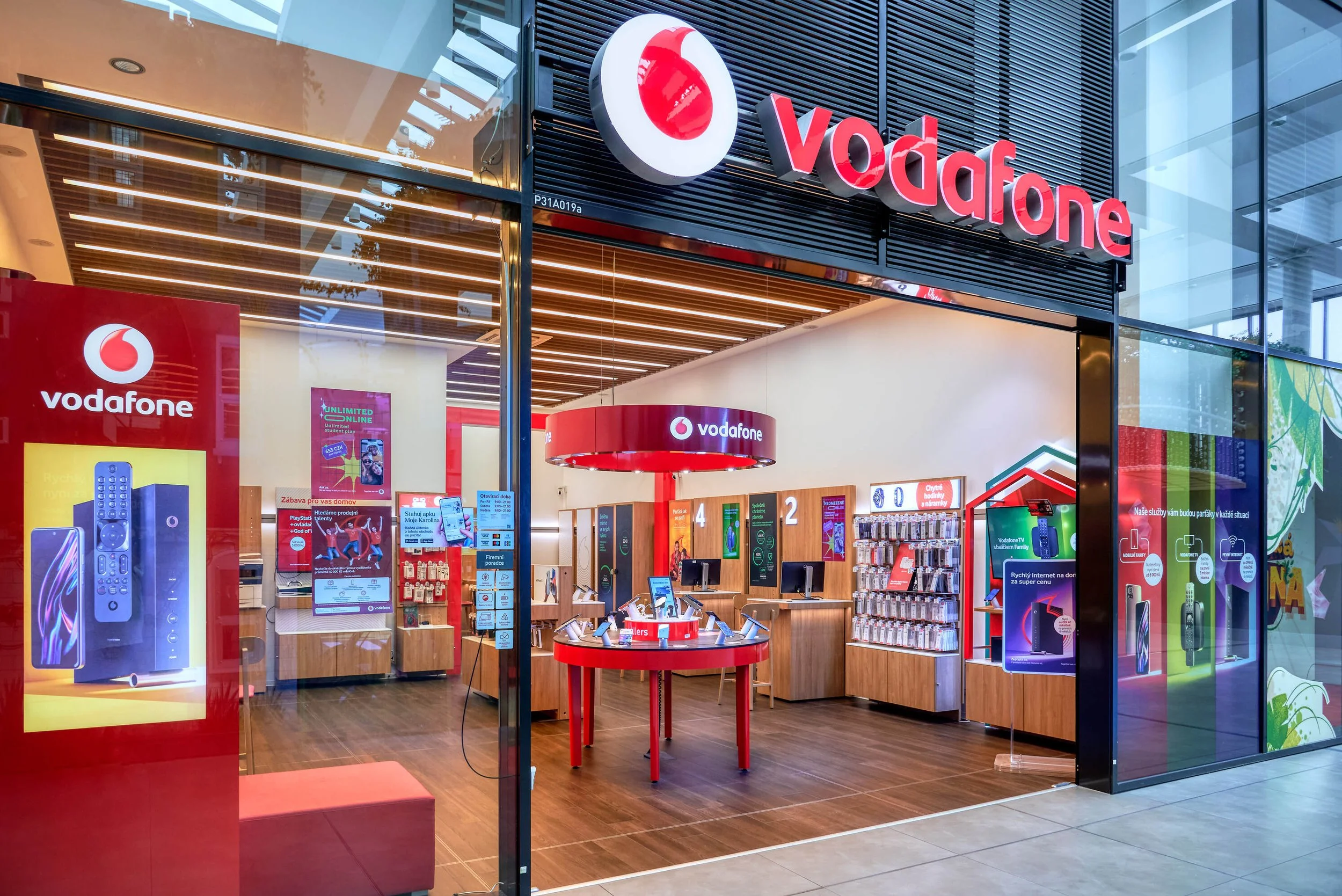 Inside a Vodafone retail store, displaying mobile phones, accessories, and promotional materials. The store has a modern design with wooden flooring, digital screens, and red accents.
