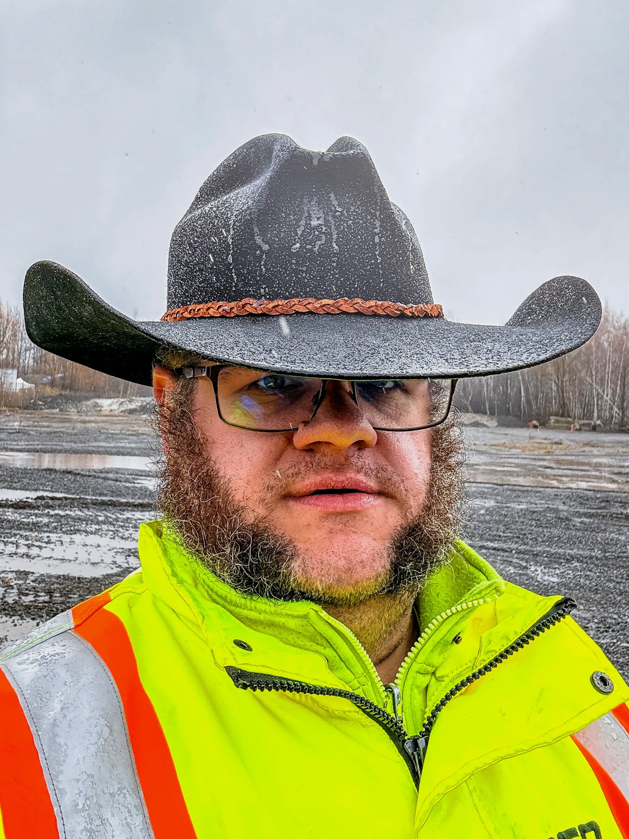 A man wearing a black cowboy hat with orange braided band, sunglasses, and a high-visibility safety jacket with yellow and orange reflective stripes stands outdoors in a snowy, overcast environment.  displaying the rugged durability of a good hat