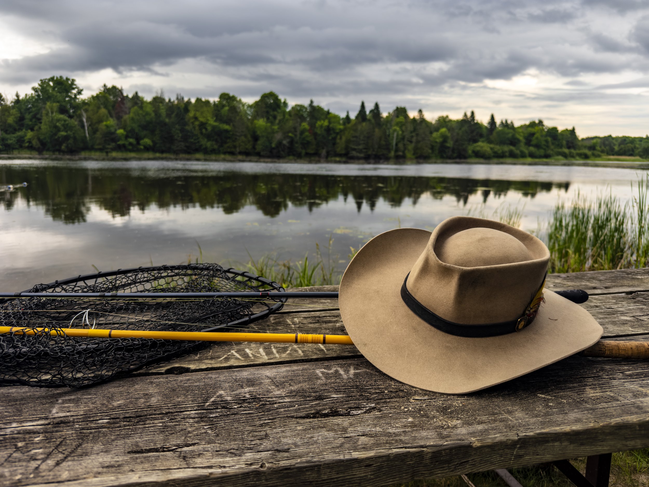 A fishing net and fishing rod lying on a wooden dock with a wide-brimmed hat resting on the edge. In the background, a peaceful lake with trees and a cloudy sky reflected on the water.