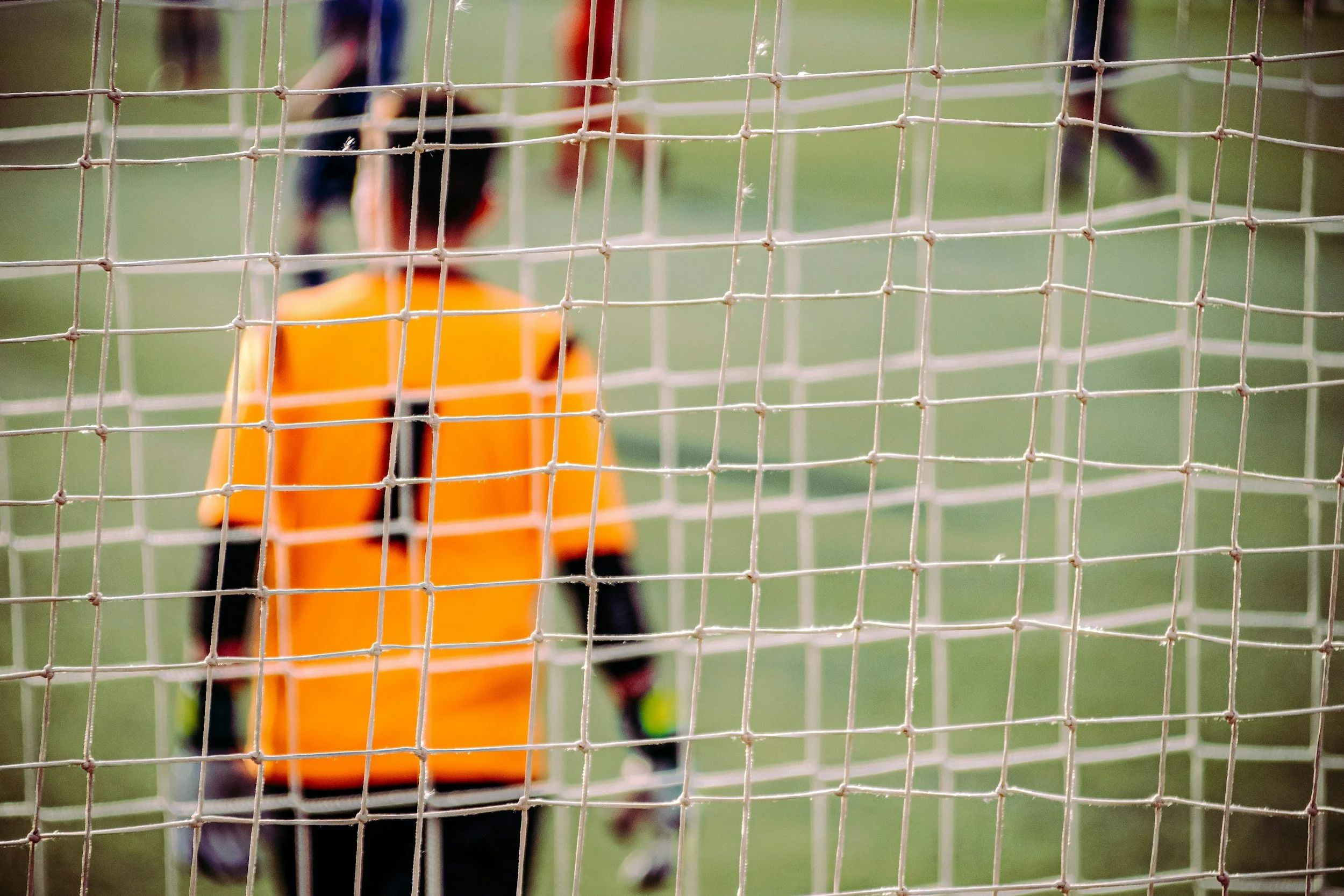 Een kind in een oranje jas die een bal in een hand vasthoudt, zichtbaar achter een gaashek op een grasveld.