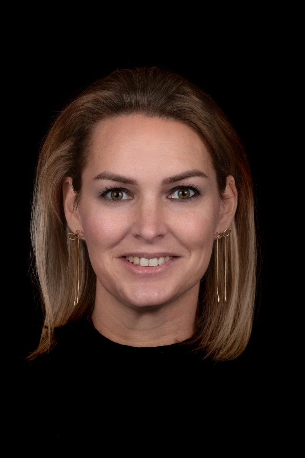 Portret van een vrouw met bruin haar, lichte huid en groene ogen, lachend met tanden zichtbaar, dragen gouden oorbellen en een zwarte top, donkere achtergrond.