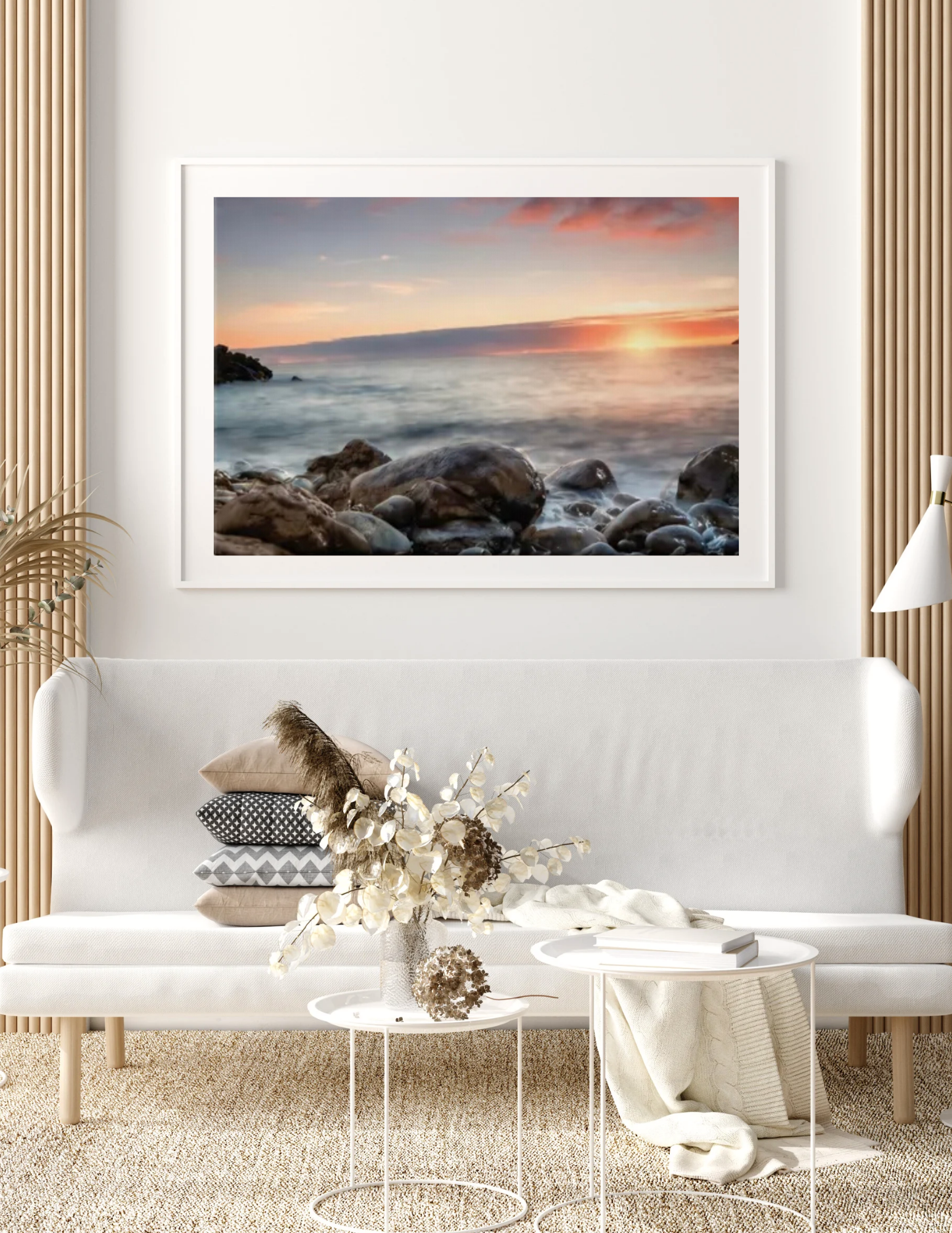 Living room with a sofa, decorative pillows, a throw blanket, a side table with a vase of dried flowers, a white coffee table with books, a large framed sunset beach photograph on the wall, beige carpet, and a floor lamp.
