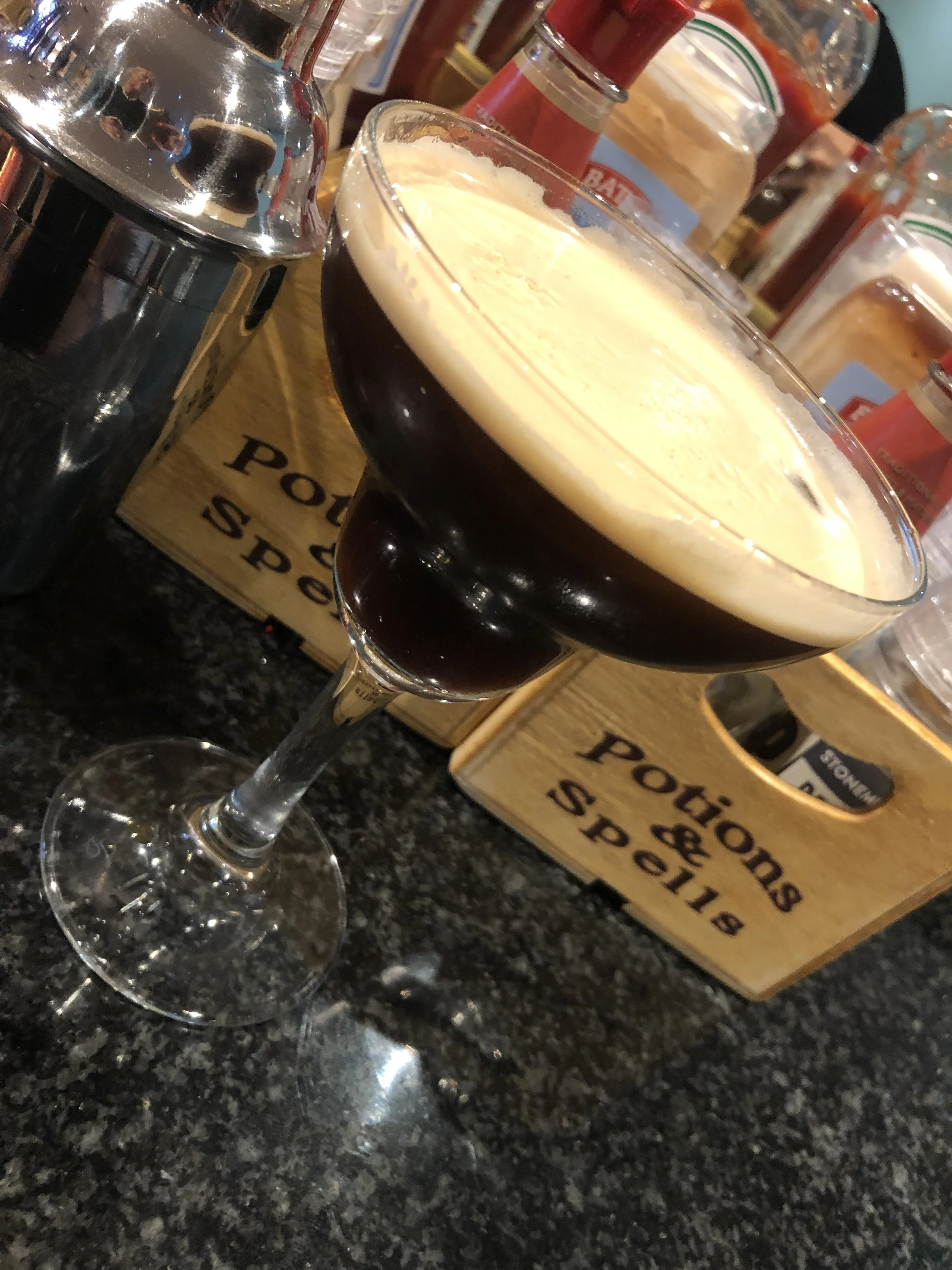 A cocktail glass filled with espresso martini topped with a frothy cream layer, placed on a dark countertop with condiment bottles and wooden crates in the background.