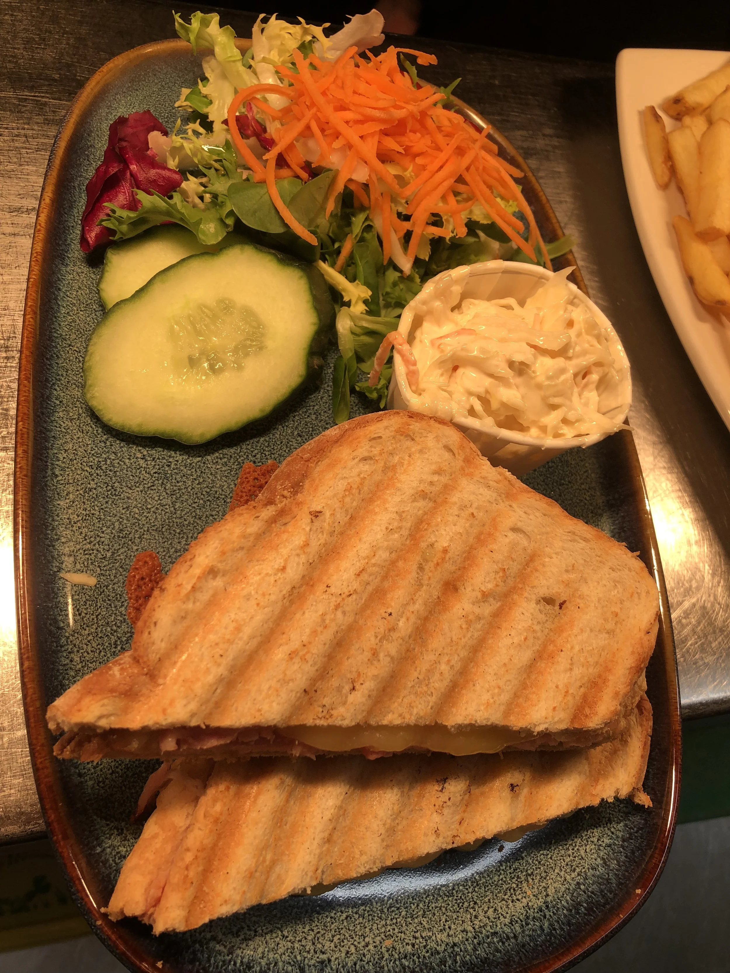 A plate with a large toasted sandwich, side salad with shredded carrots, lettuce, and cucumber slices, a small cup of coleslaw, and a portion of potatoes.
