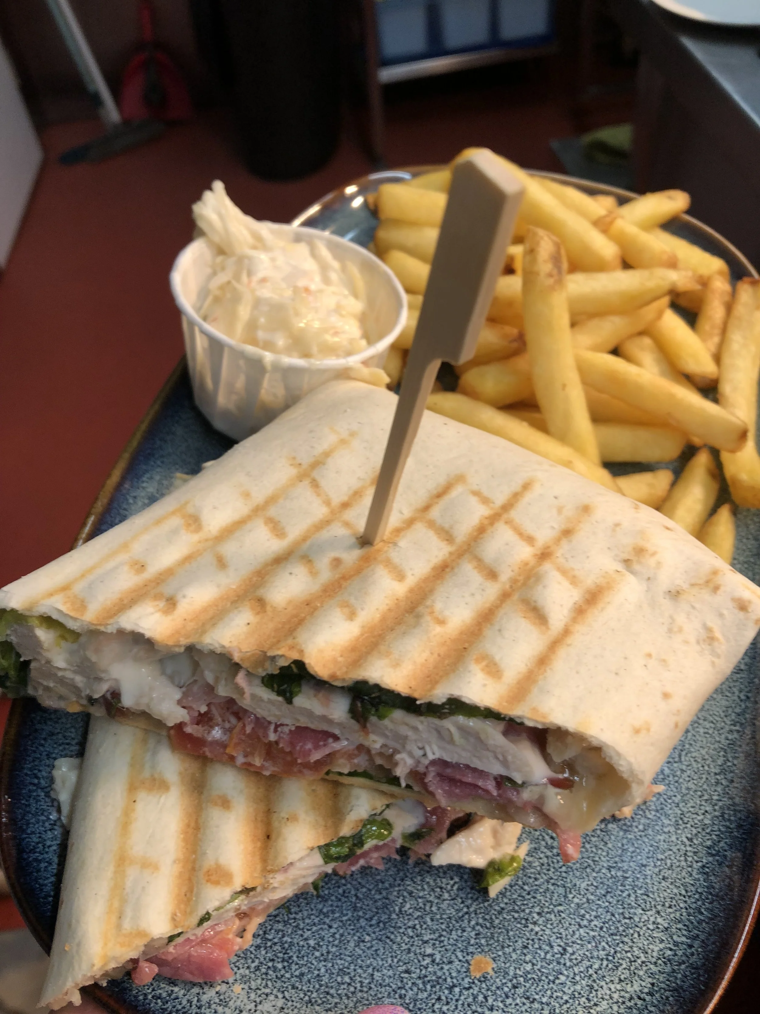 A panini sandwich with grilled bread, filled with meats, cheese, and vegetables, along with a side of French fries and a small container of potato salad.