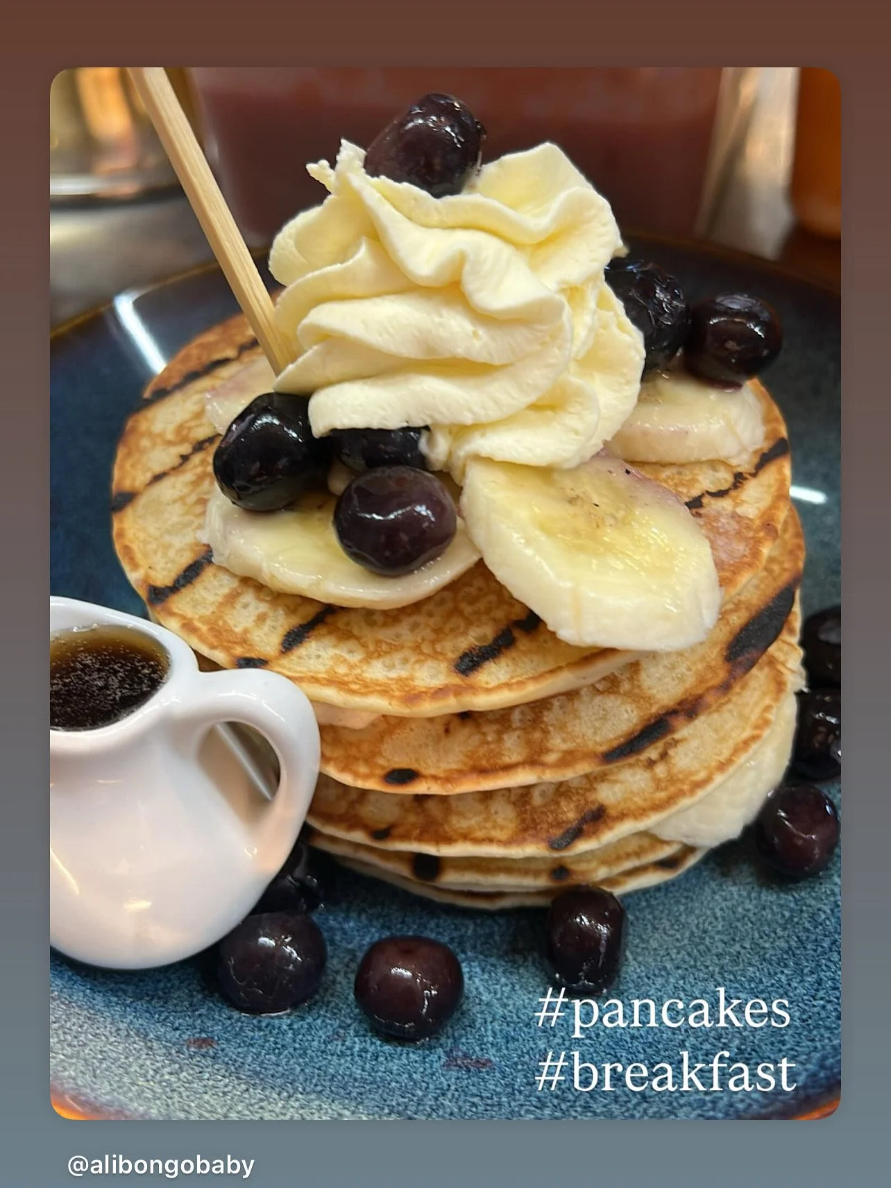 Stack of three grilled pancakes topped with banana slices, blueberries, and whipped cream, with syrup in a small pitcher, on a blue plate, with hashtags #pancakes and #breakfast.