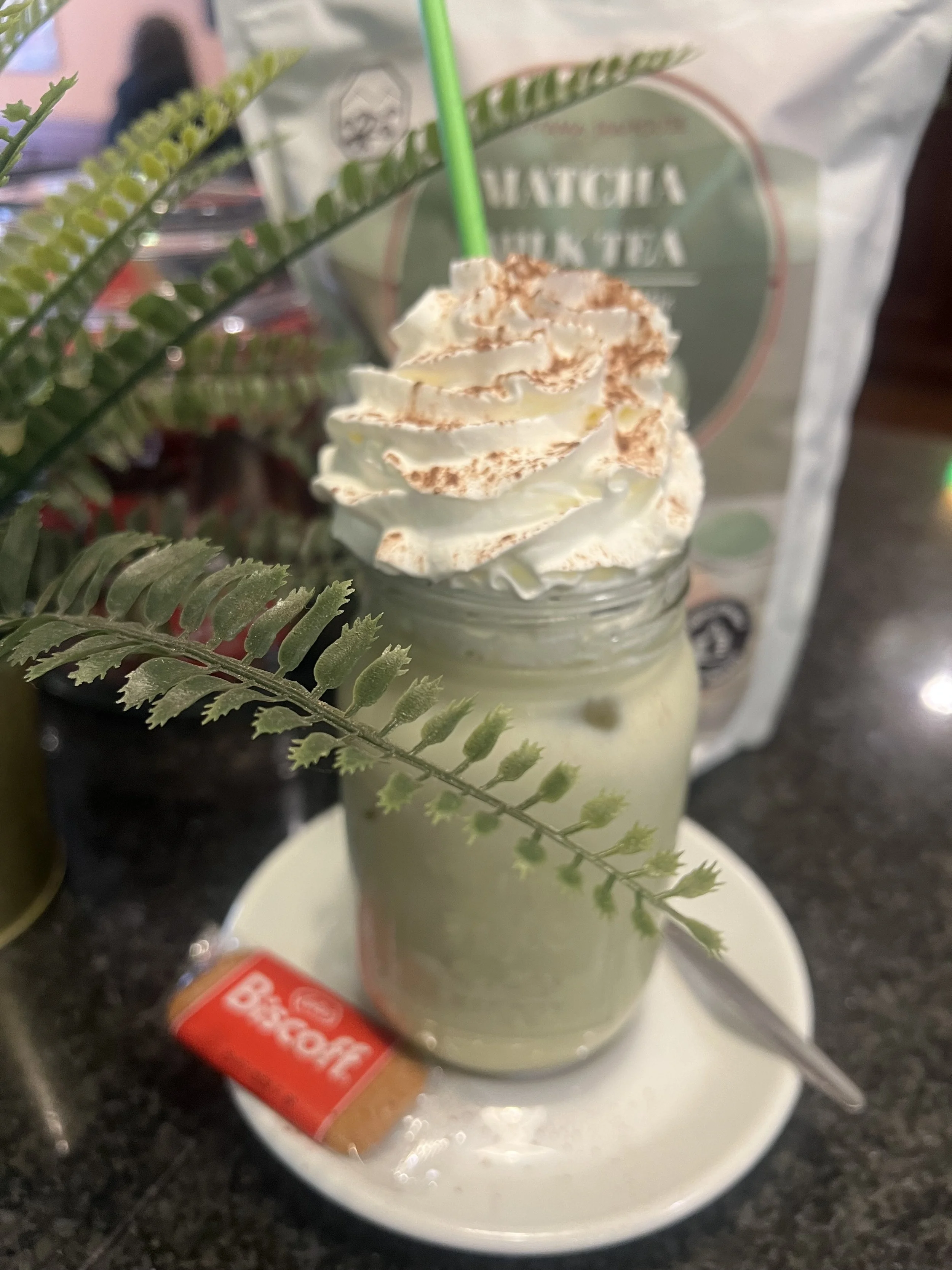 A drink with whipped cream topped with cocoa powder inside a glass jar with a green straw, placed on a white plate in front of a bag of matcha tea, with green fern leaves decoration in the foreground.