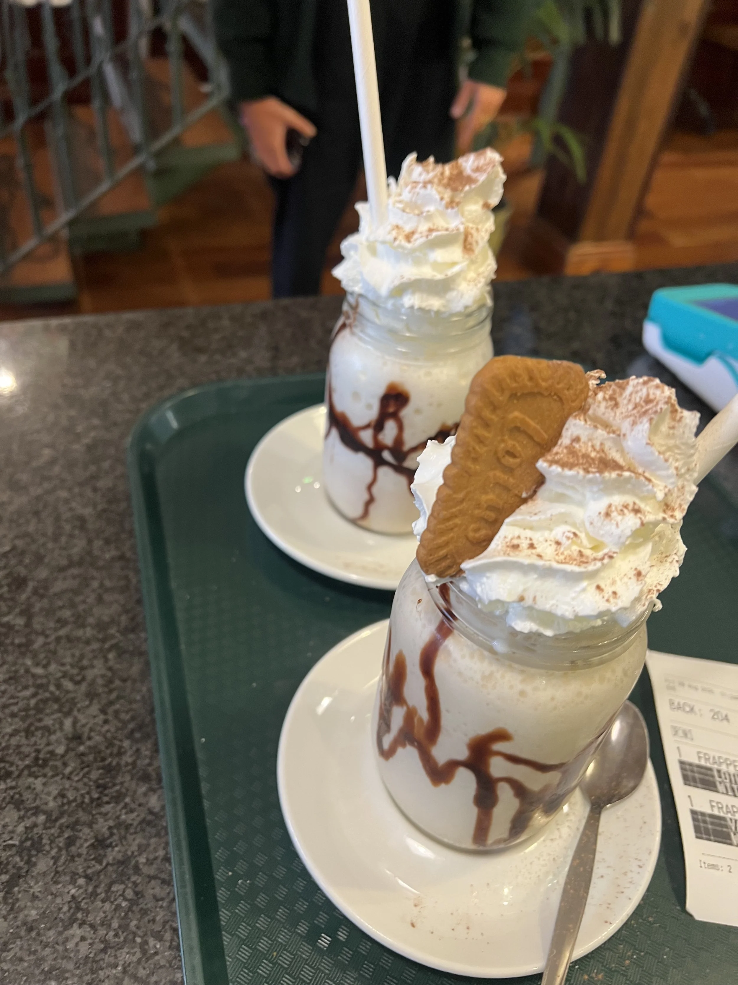 Two milkshakes topped with whipped cream, sprinkled with cocoa powder, and garnished with a cookie, served on white plates on a green tray at a restaurant.