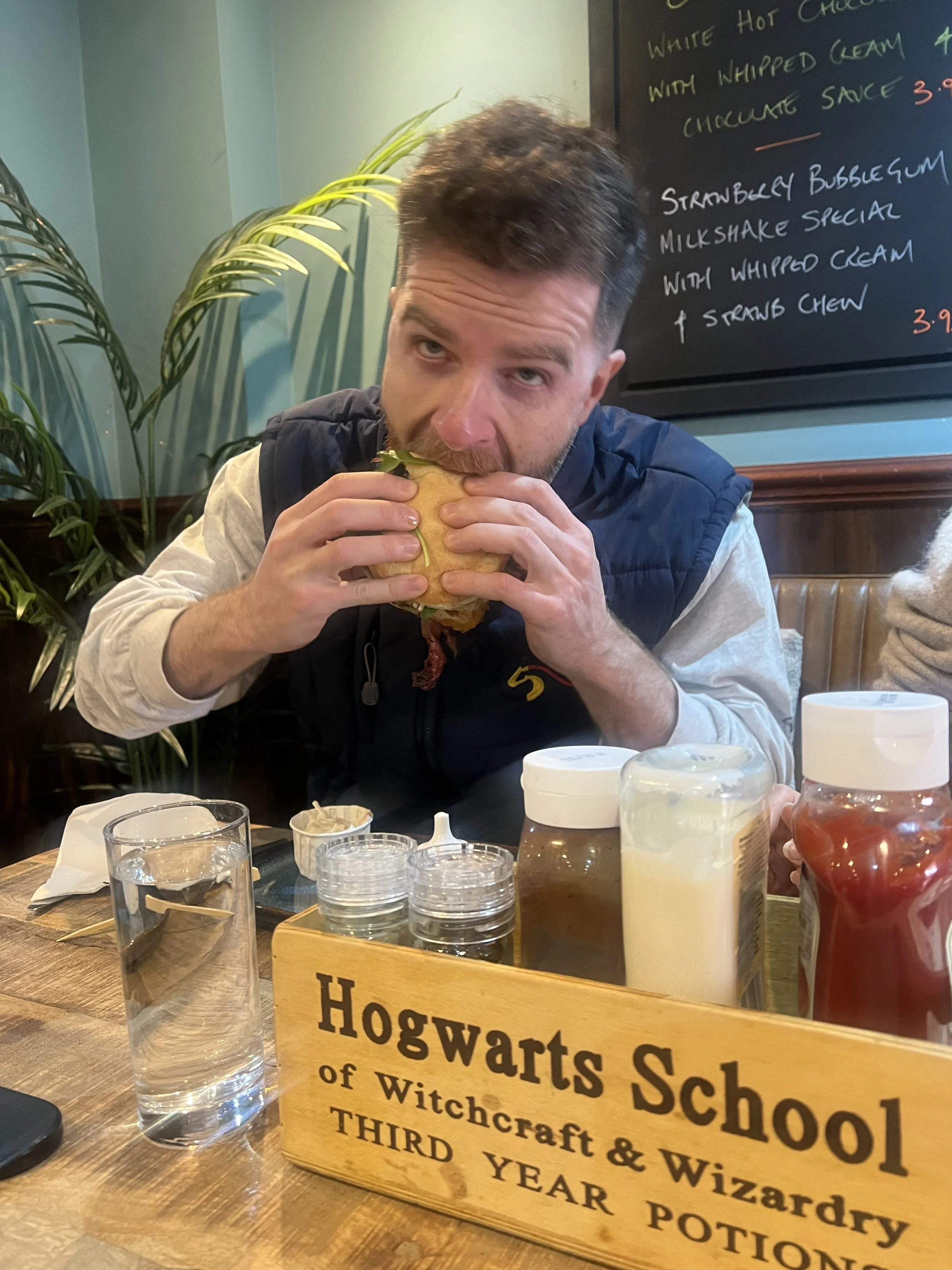 A man with short dark hair and a beard is sitting at a wooden table in a restaurant, holding and about to eat a sandwich. In the background, there is a blackboard with a handwritten menu, some houseplants, and various drink glasses and condiment jars