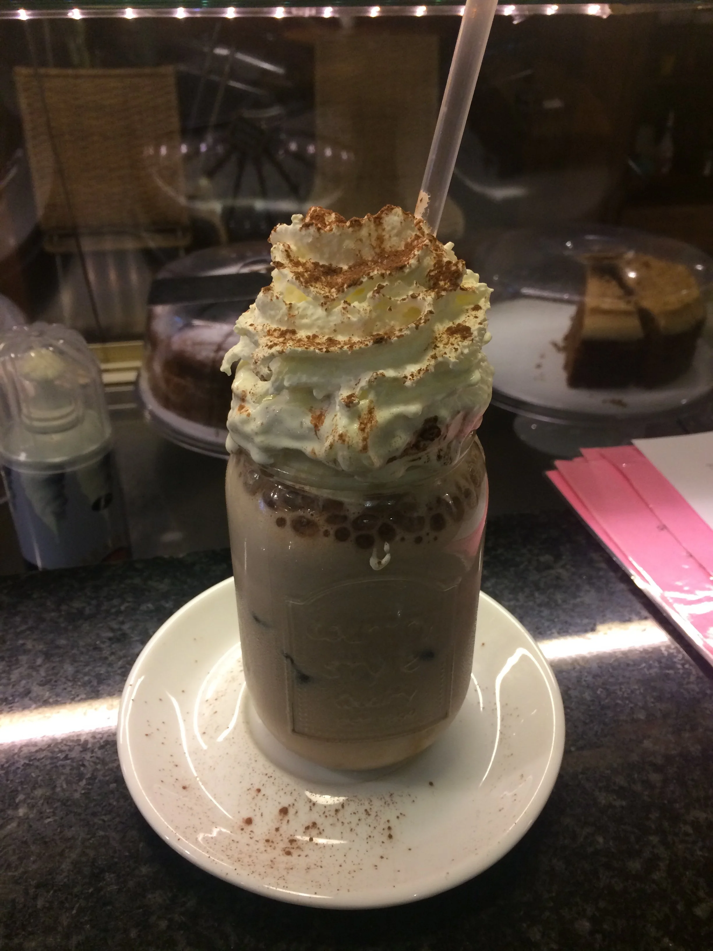 A tall glass of chocolate beverage topped with whipped cream and cocoa powder, served on a white oval plate with some cocoa powder sprinkles around the outside.