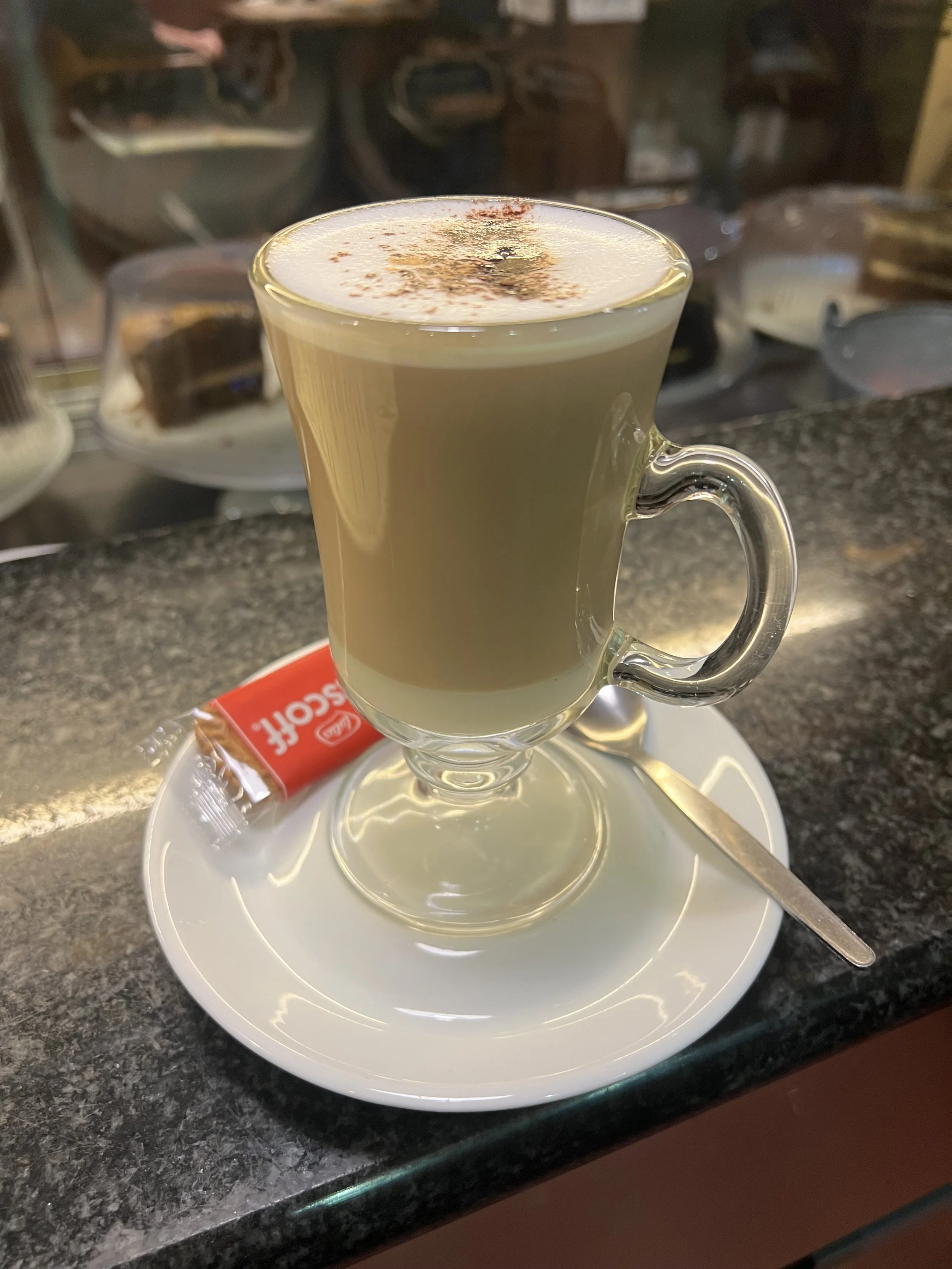 A clear glass mug filled with a layered coffee beverage topped with whipped cream and sprinkled cinnamon, placed on a white saucer with a packaged sugar or sweetener. The mug is on a dark countertop with a blurred background of various dishes and kit