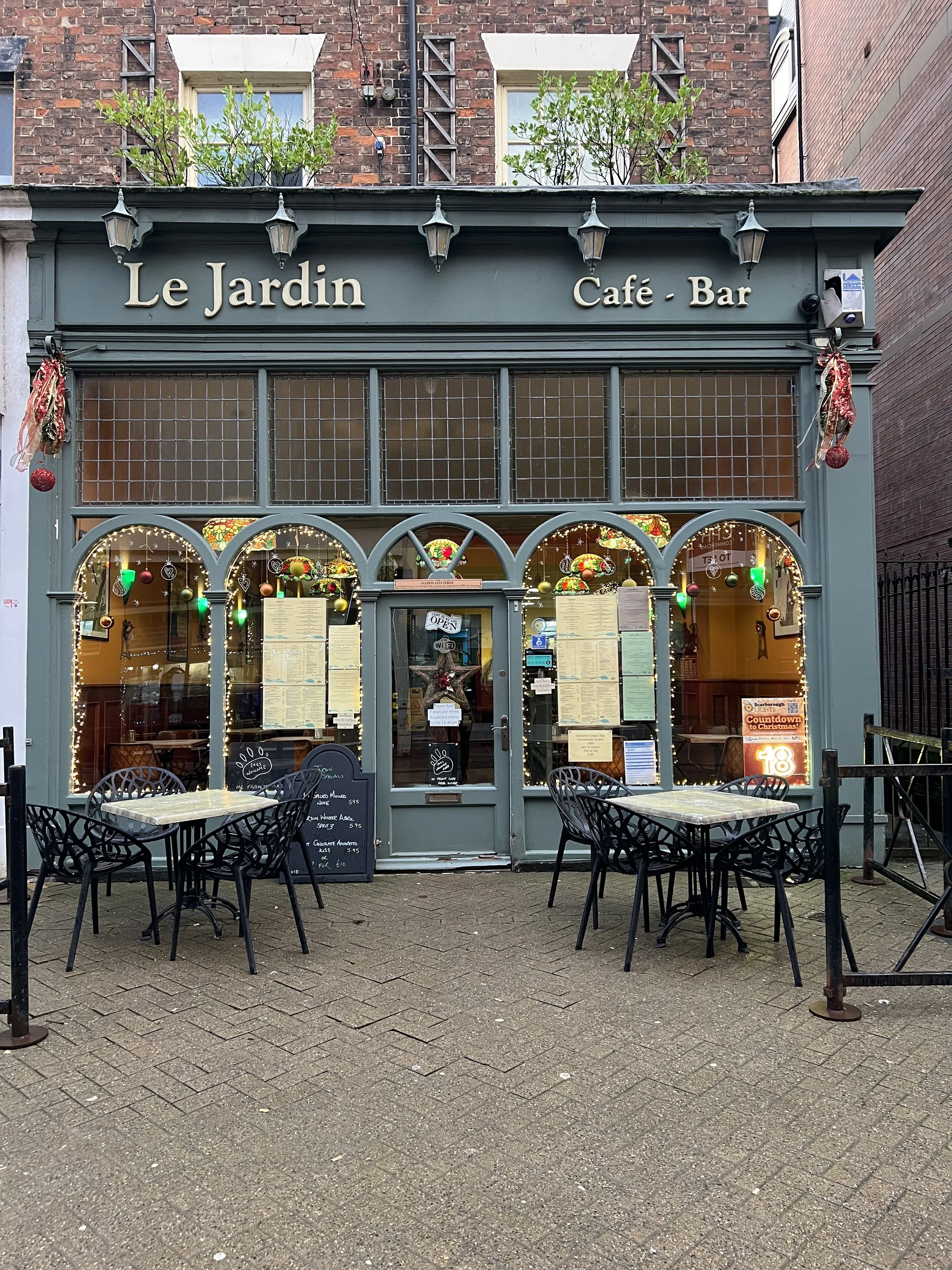 The exterior of a small café named 'Le Jardin,' decorated with Christmas ornaments and lights, with outdoor seating consisting of three round tables and several chairs, and a chalkboard menu in front.