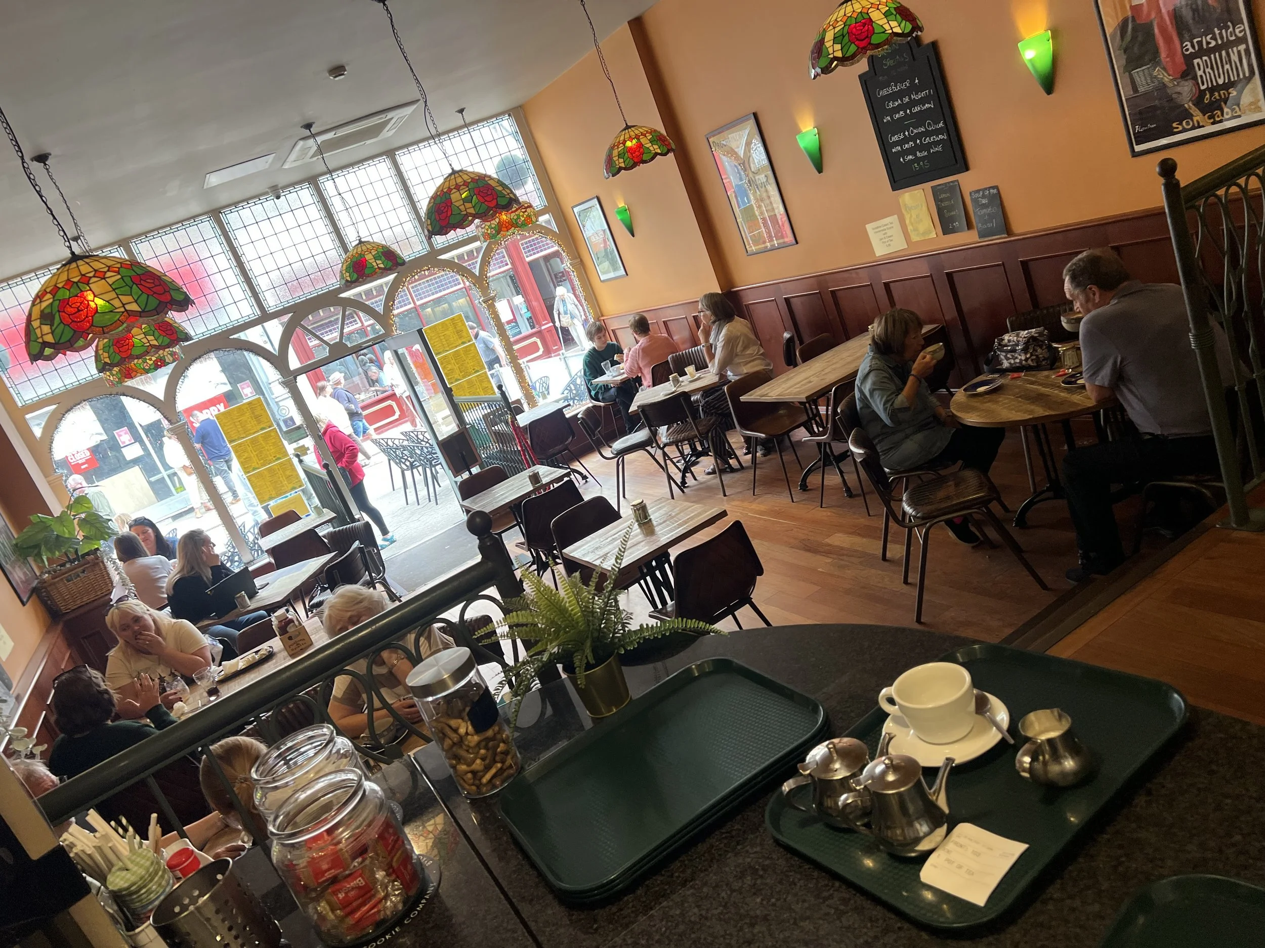 Inside a cozy restaurant with colorful stained glass hanging lamps, wooden paneling, and framed artwork. Customers are seated at tables, enjoying meals and drinks, with a view outside through large windows.