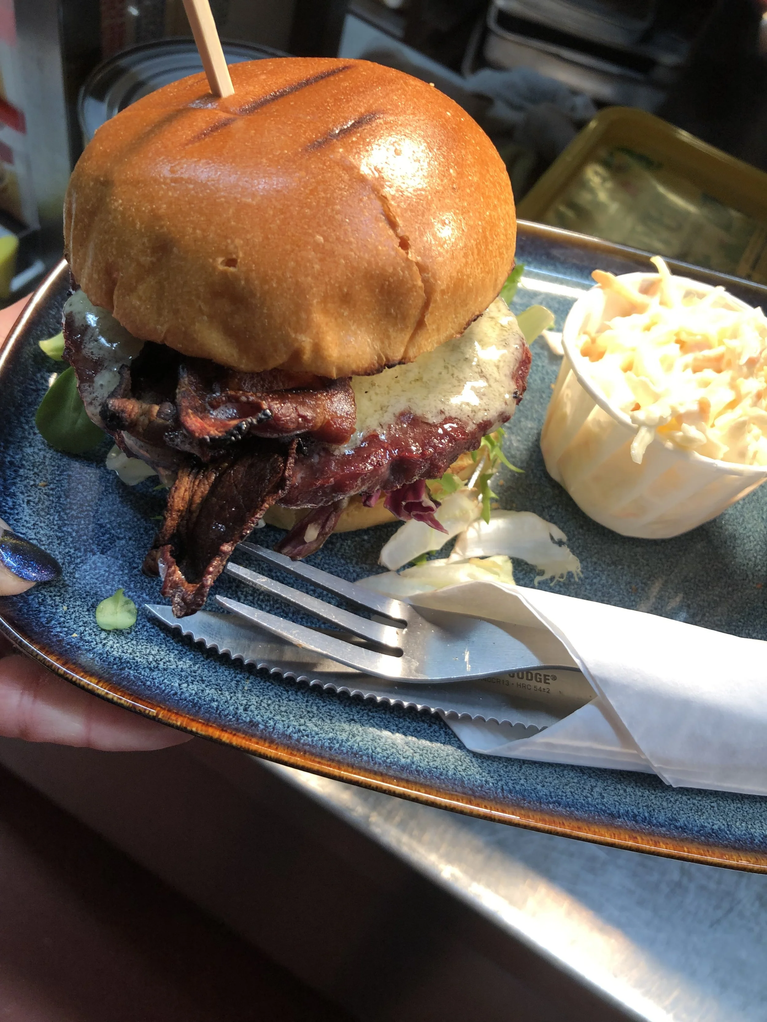 A cheeseburger with lettuce, tomato, bacon, cheese, and a bun, served with coleslaw on a metal tray.