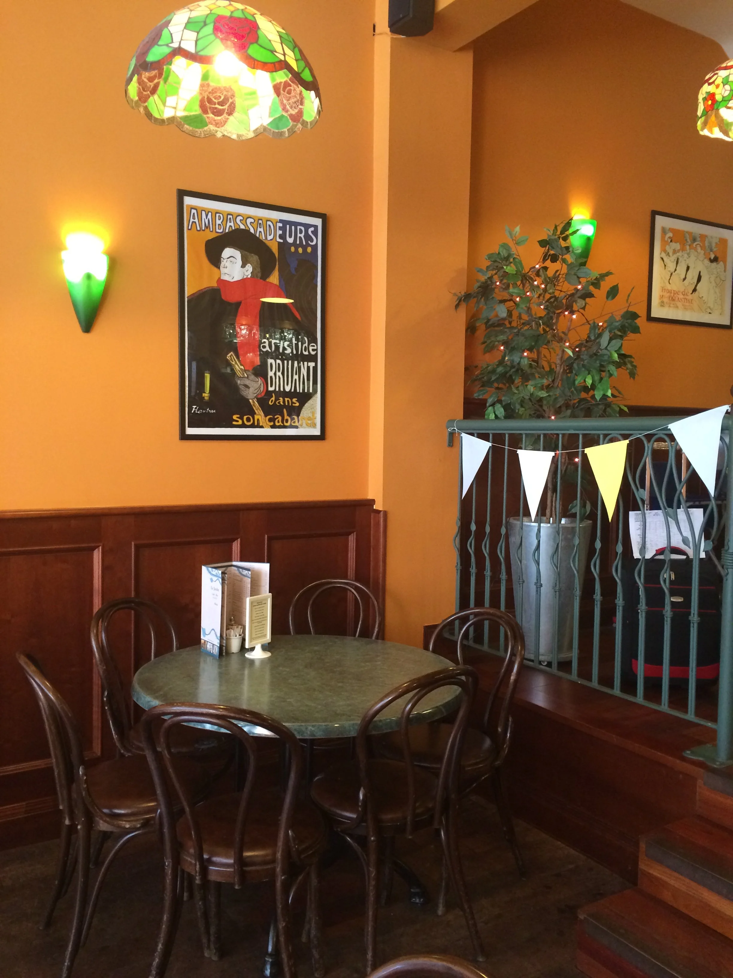 A cozy indoor restaurant scene with a round wooden table surrounded by six wooden chairs, decorated with menus and small containers, with warm orange walls, colorful stained glass hanging lamps, a potted plant, framed artwork, and a garland of yellow