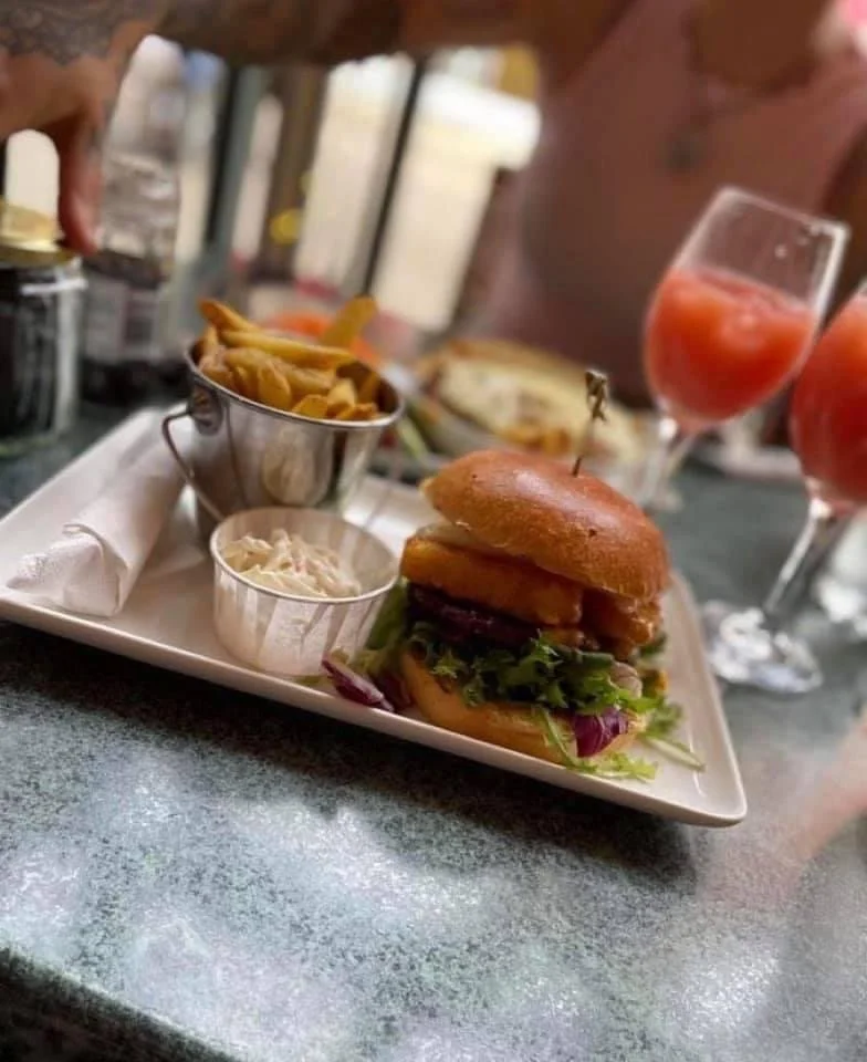 Burger with lettuce, tomato, and fried onion rings, served with french fries and coleslaw on a white plate, with two glasses of cocktails in the background.