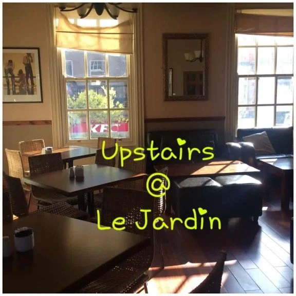 Inside a cozy cafe, with sunlight streaming through large windows, tables and chairs arranged around the room, a mirror on the wall reflects interior details, and handwritten yellow text reads 'Upstairs @ Le Jardin'.