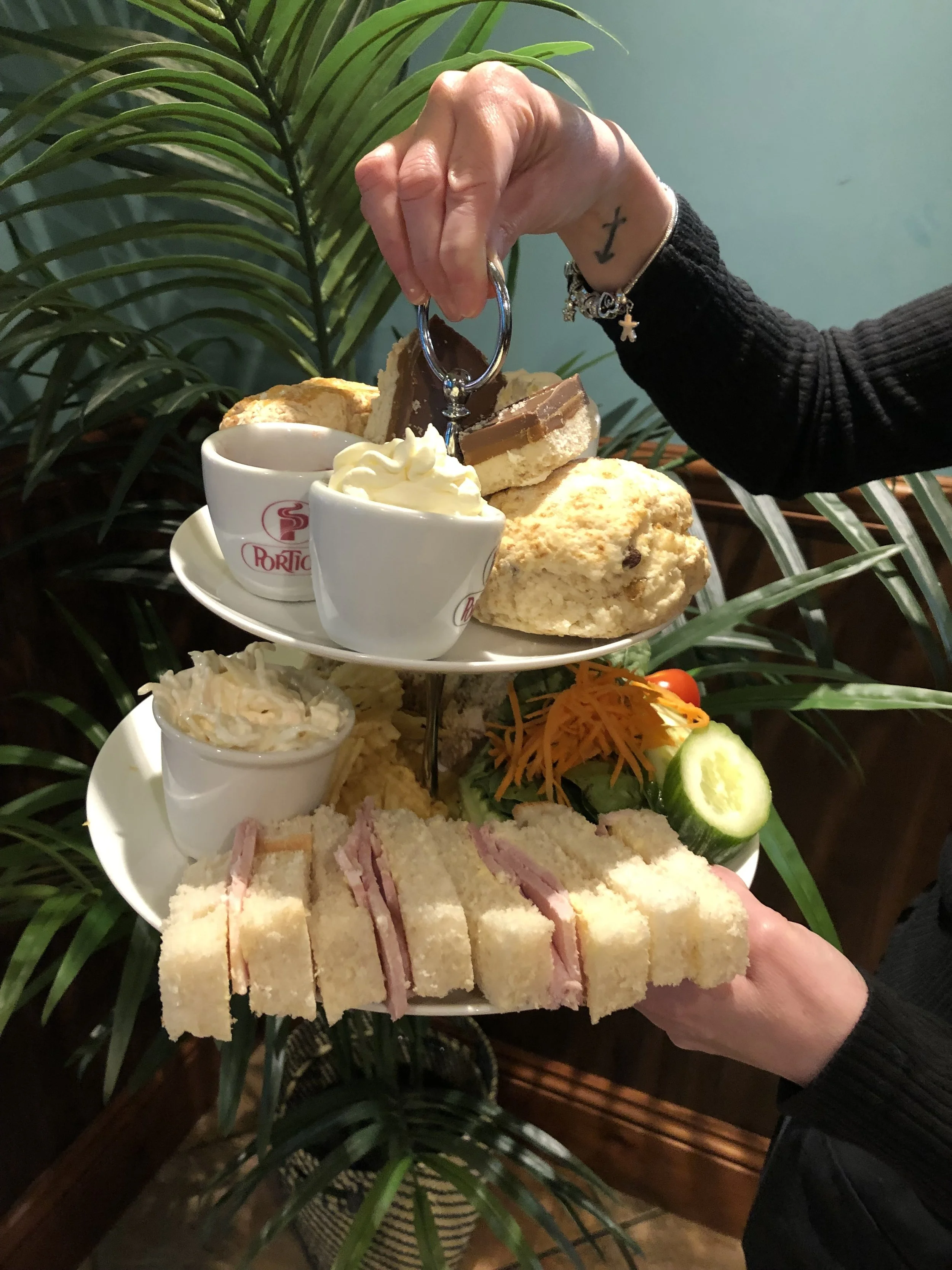 A two-tiered tray of assorted food items including sandwiches, cakes, a small cup of coleslaw, whipped cream, and condiments, with a person holding a finger sandwich in front of a green leafy plant and blue wall.