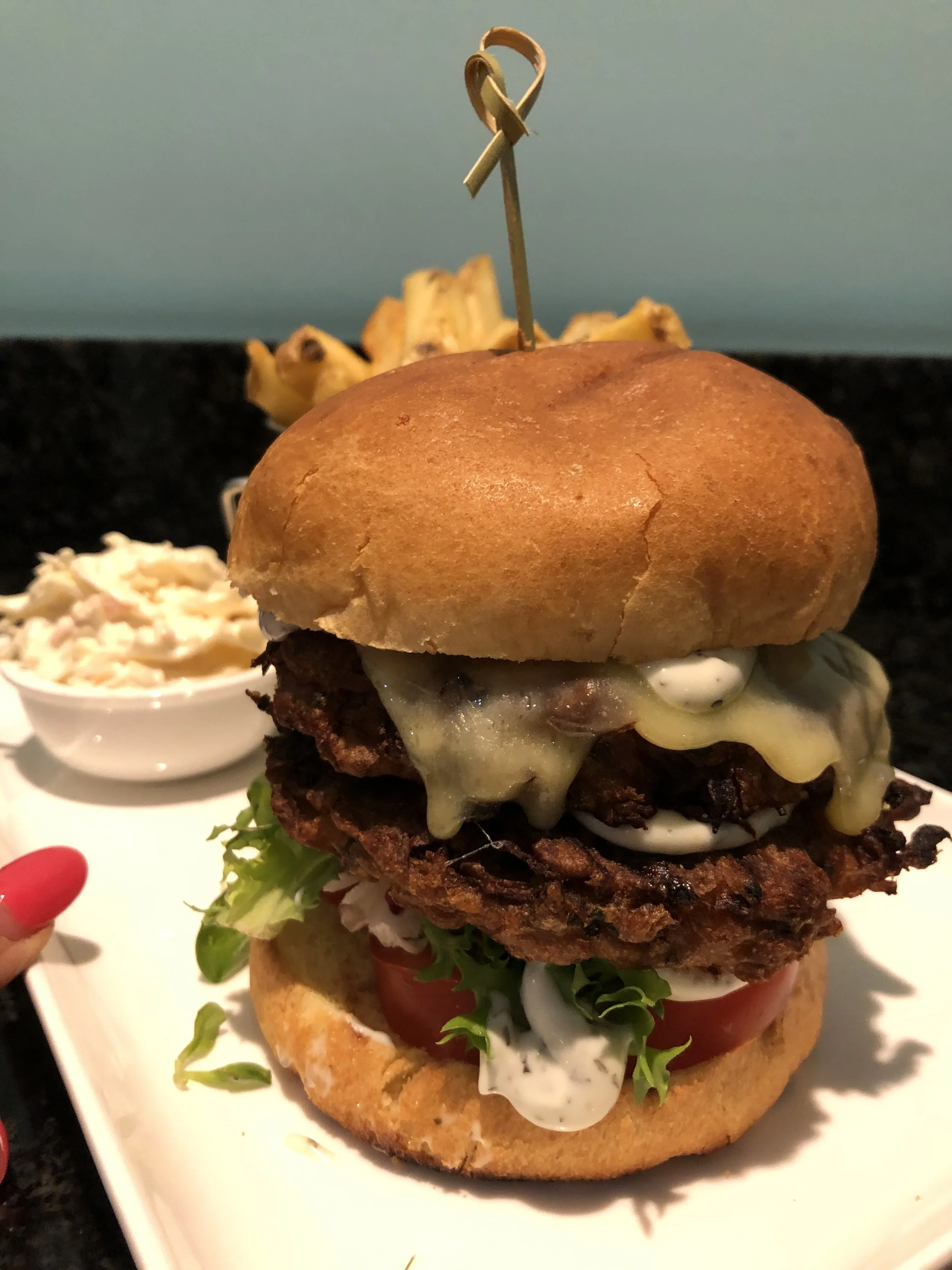 A close-up of a gourmet burger with lettuce, tomato, cheese, grilled beef patty, fried onion ring, and a bun with a decorative skewer on top, served on a white plate with a side of coleslaw.