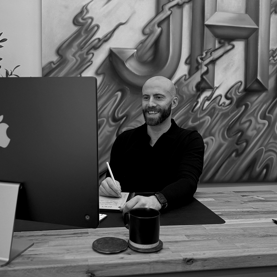 A man with a beard and a bald head, smiling at his computer in a modern office space, with an abstract mural on the wall behind him.