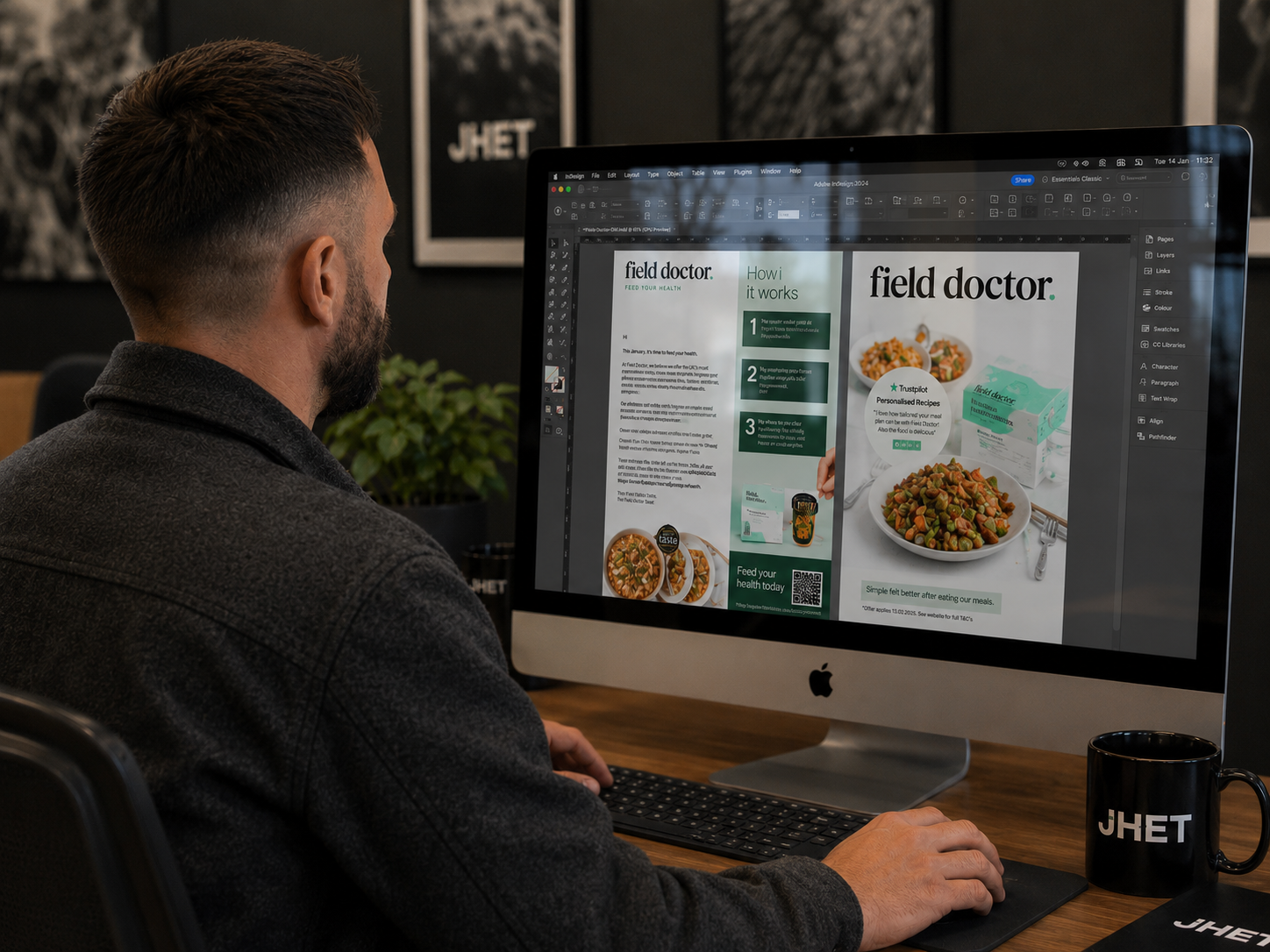 A man working on a graphics design on an iMac computer in an office. The screen displays a flyer or presentation about a product called 'field doctor'. The desk also has a black mug with the logo 'JHET' and a potted plant in the background.