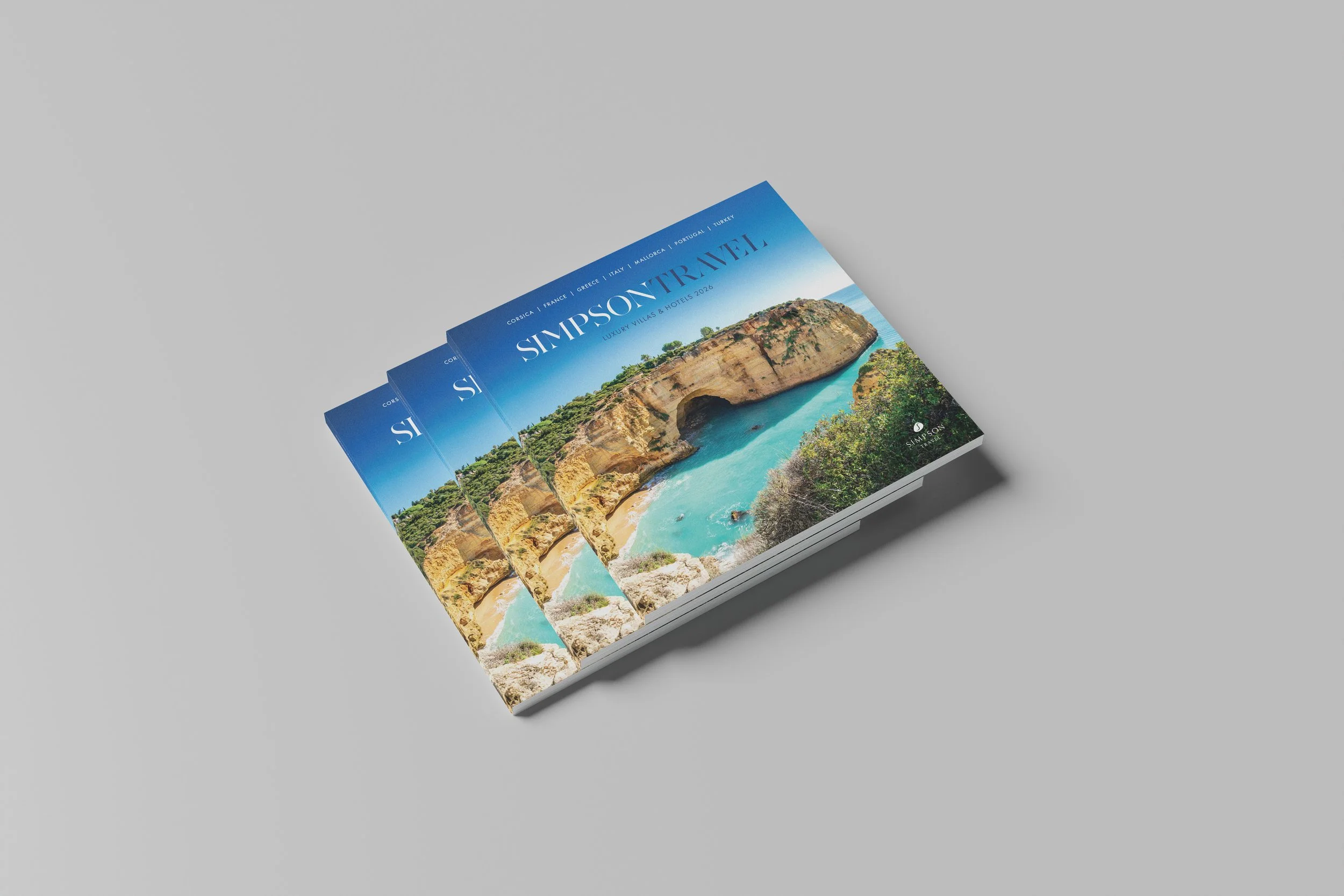 Stack of brochures featuring a scenic coastal landscape with cliffs and ocean, titled 'SIMPSONTRAVEL' highlighting luxury villas and hotels in Corsica, France, Greece, Italy, Mallorca, Portugal, and Turkey.