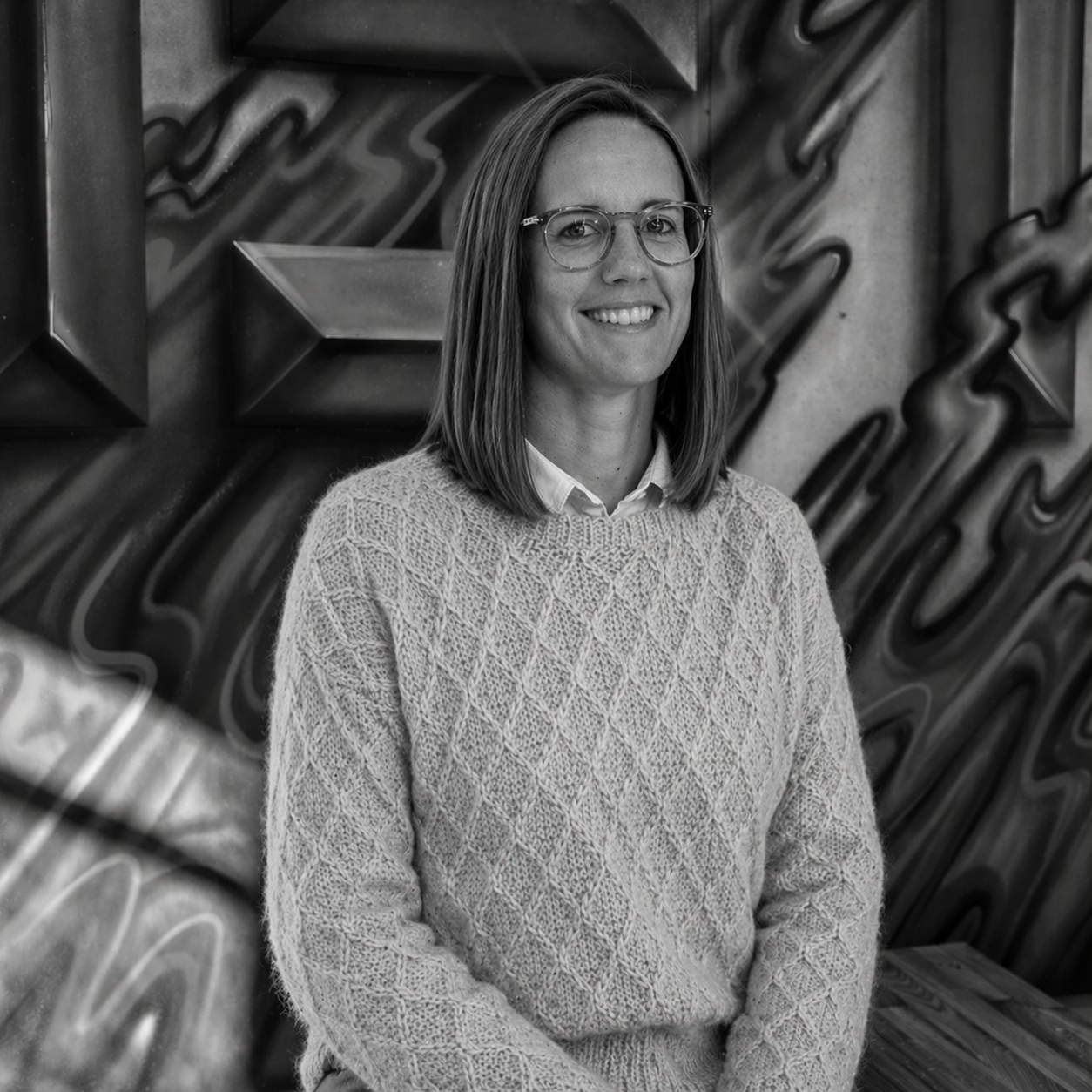 A woman with shoulder-length hair wearing glasses, a collared shirt, and a knit sweater, smiling while standing in front of a textured wall with geometric and abstract patterns.
