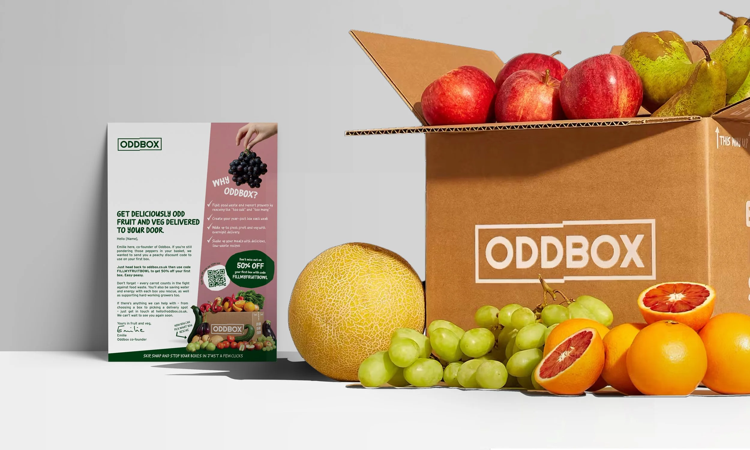 Assorted fresh fruits including apples, pears, grapes, oranges, and a halved grapefruit, alongside a brown cardboard box labeled 'ODDBOX' and a flyer promoting organic fruit and vegetable delivery.