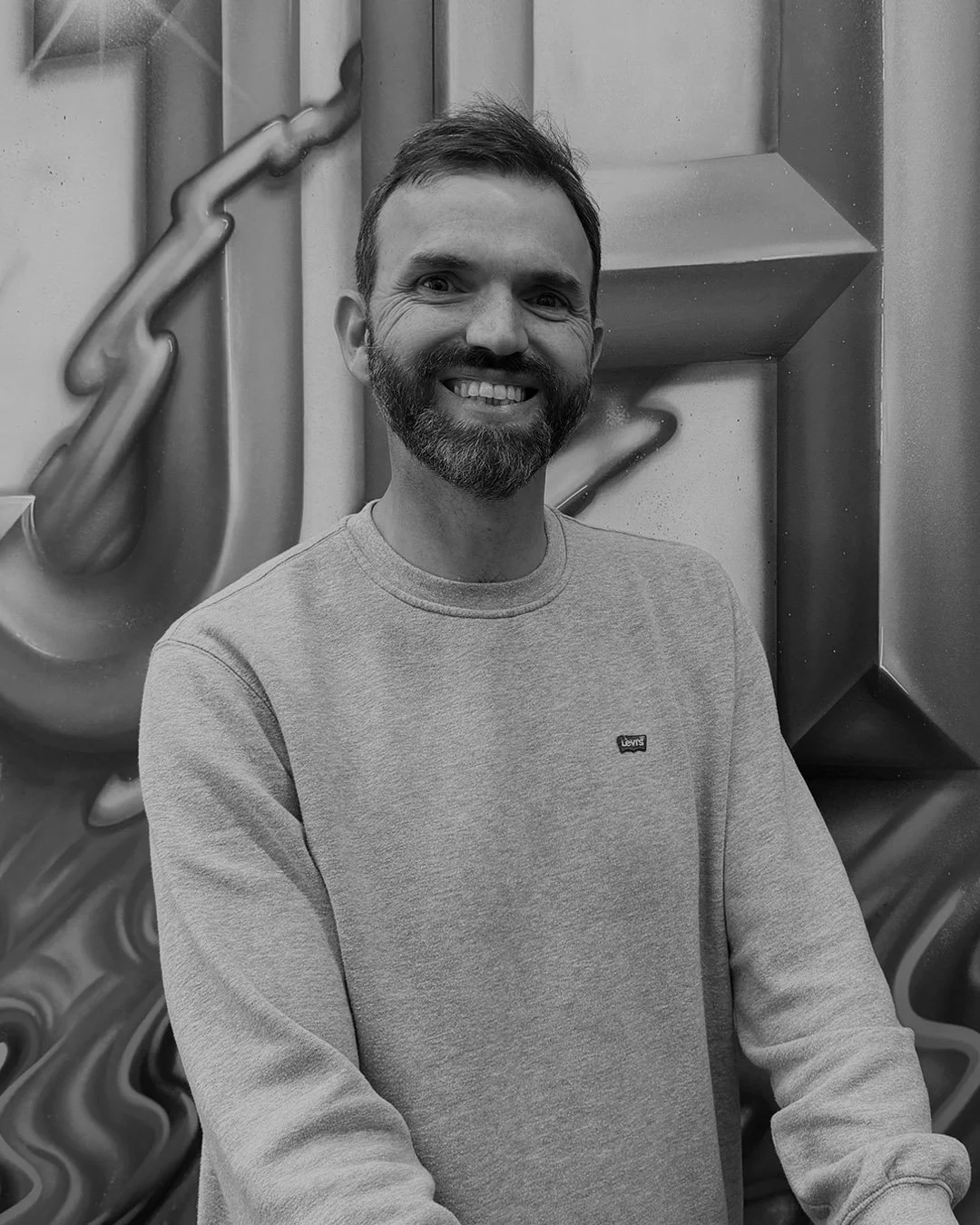 A smiling man with a beard and short hair standing in front of an abstract metallic background.