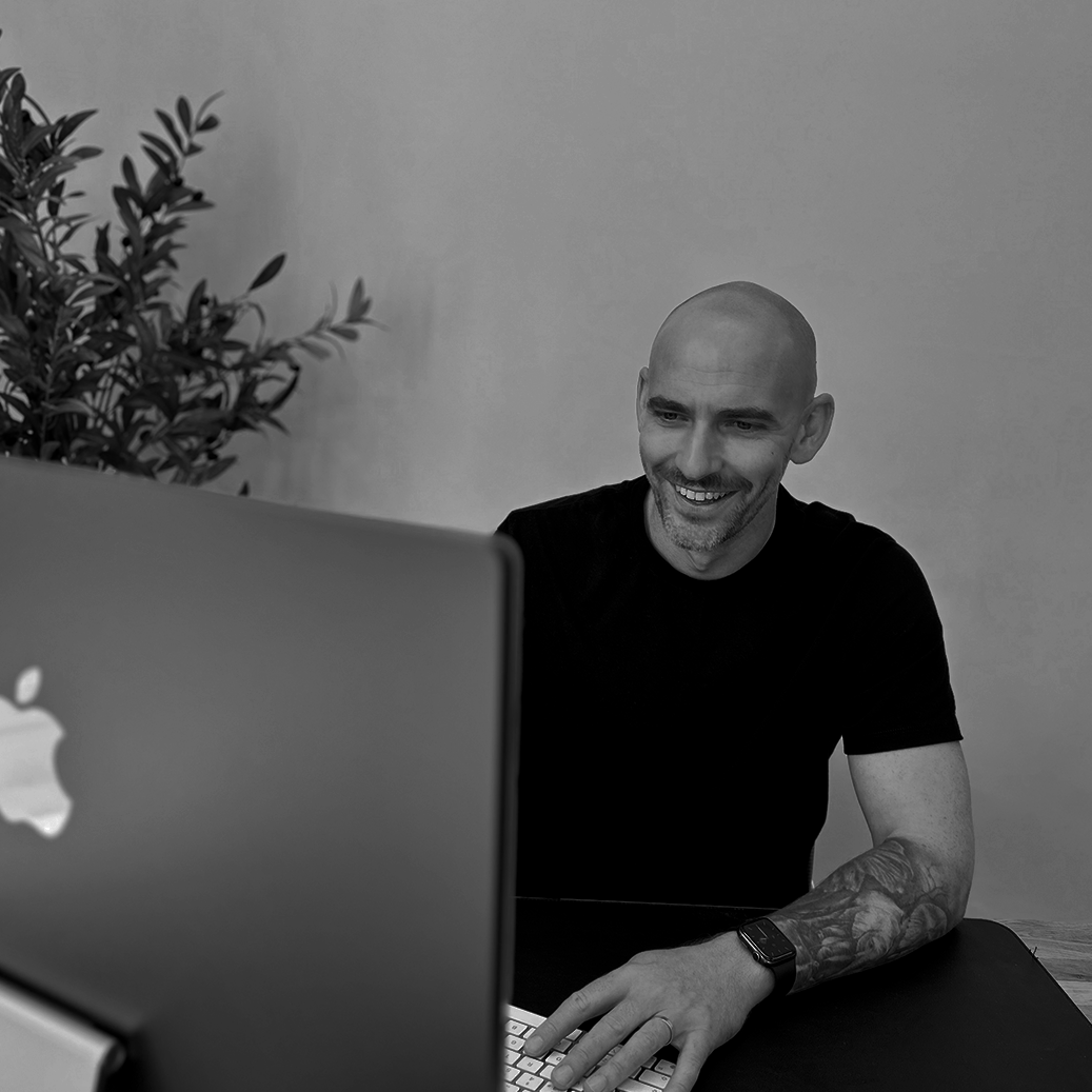 Man with a tattooed arm buttoning a computer in front of him, smiling while looking at the screen, with a plant in the background.