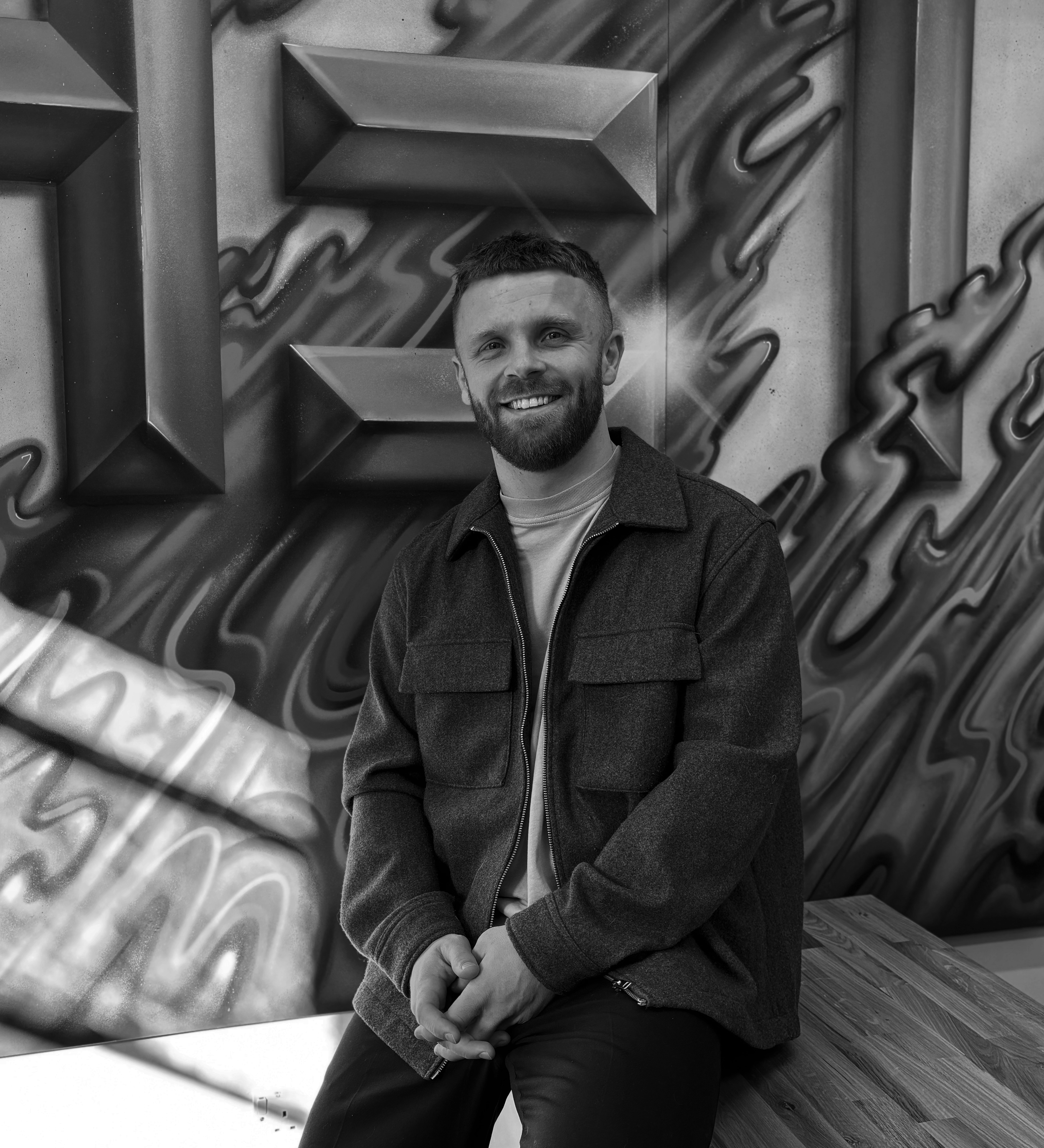 Black and white photo of a man with a beard and short hair, smiling, seated with his hands clasped. He is wearing a dark jacket and a light-colored shirt, against an abstract textured wall with wood flooring visible at the bottom.