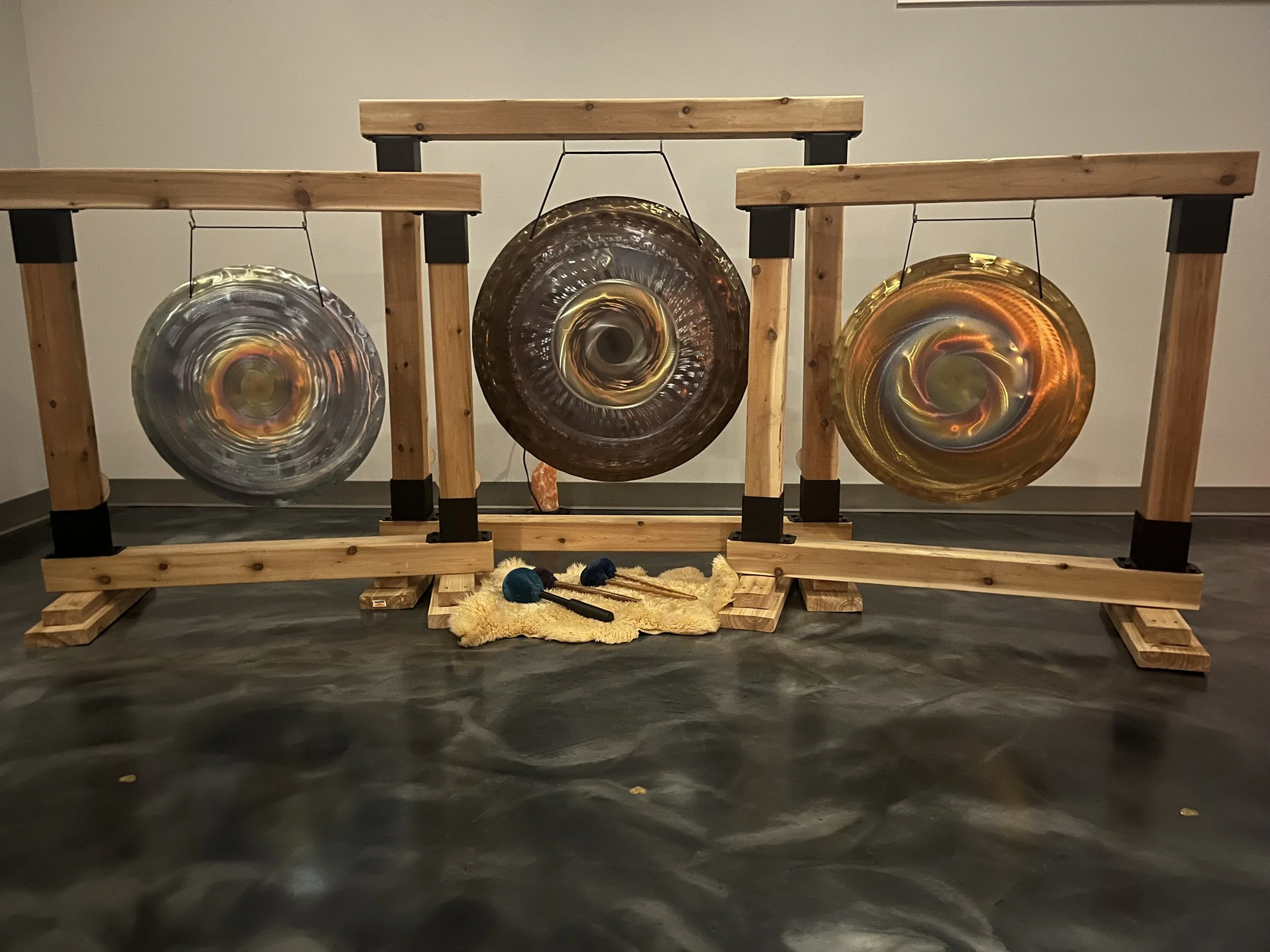 Three large circular metal gong drums hanging on a wooden frame, with mallets resting below on a yellow cloth, in an indoor setting.