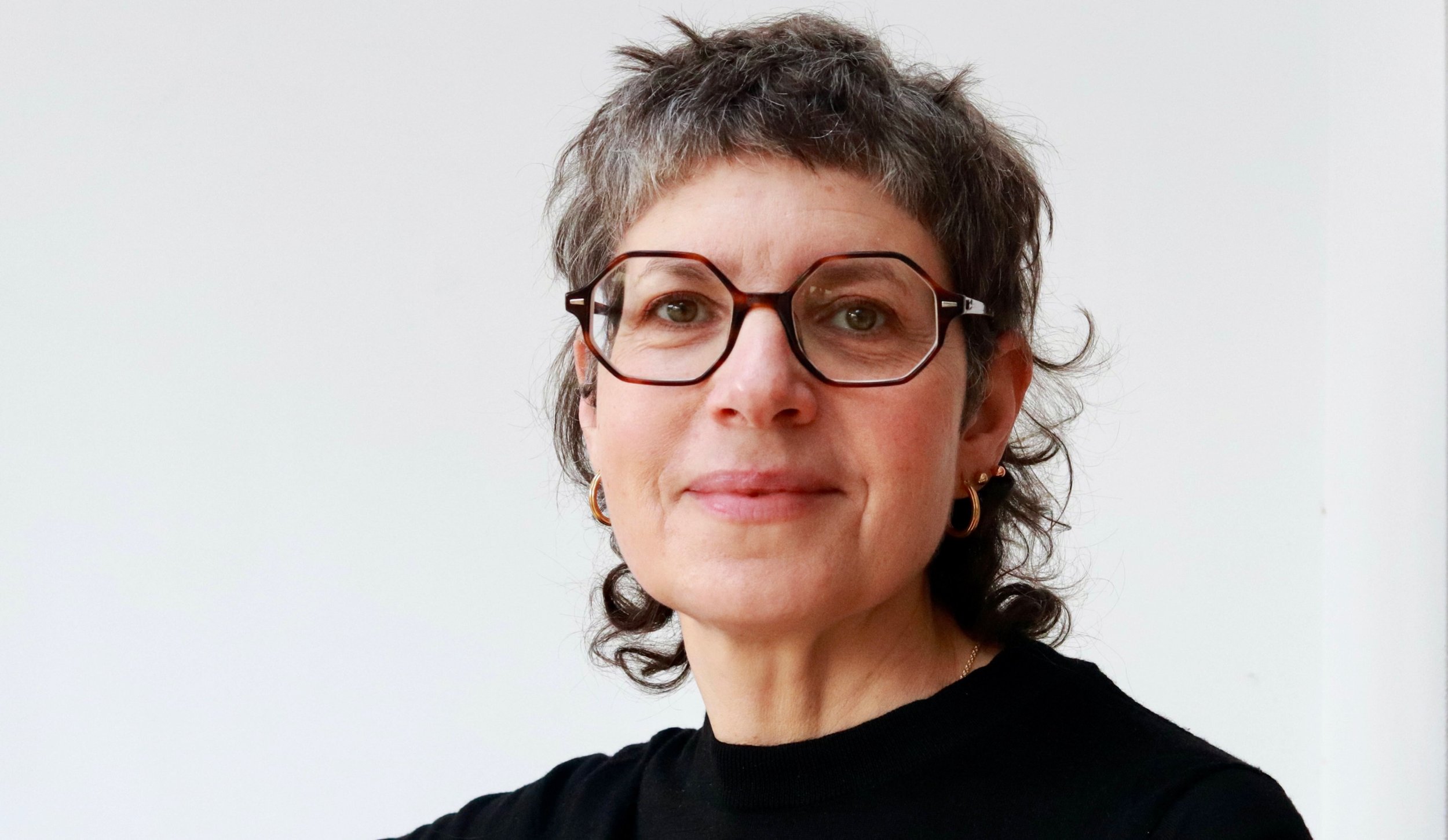 Close-up portrait of a Sonia Boué with short, curly brown hair, wearing large, geometric glasses, against a plain white background.