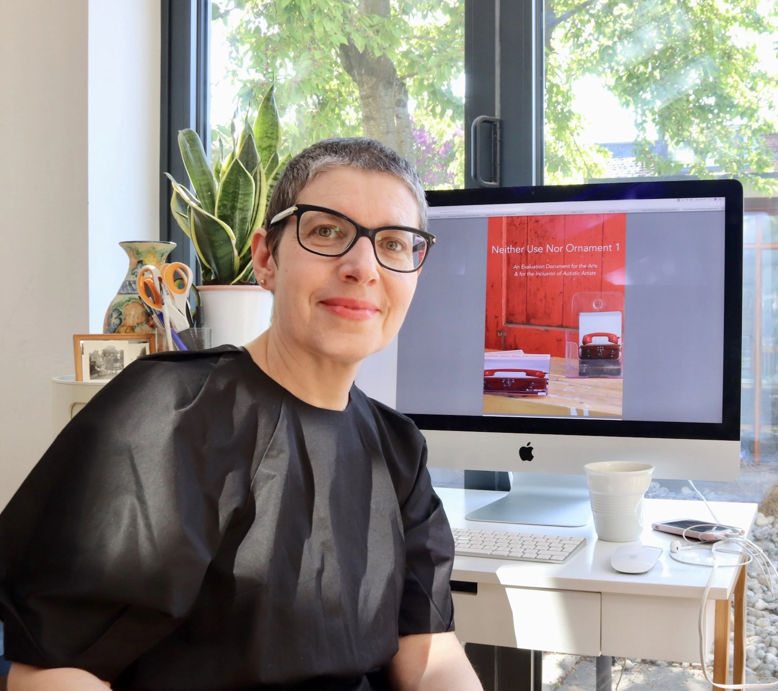 A woman wearing glasses sitting at a desk in an office with a computer, houseplants, and a window showing greenery outside.