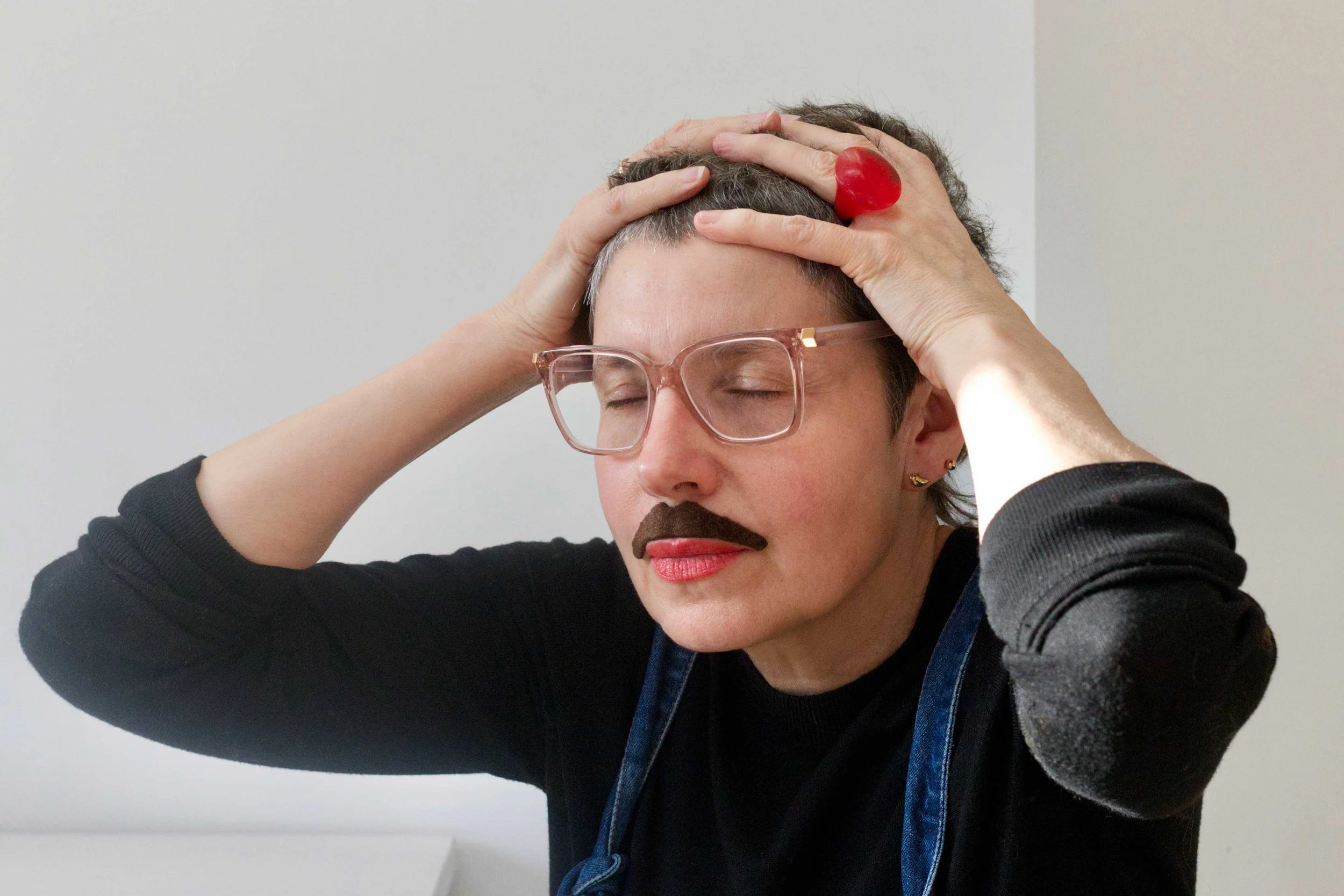 Portrait of Sonia Boué. A woman with short dark hair, wearing glasses and a false moustache, holding their head with both hands, wearing a black top and a blue apron in front of a plain white wall.