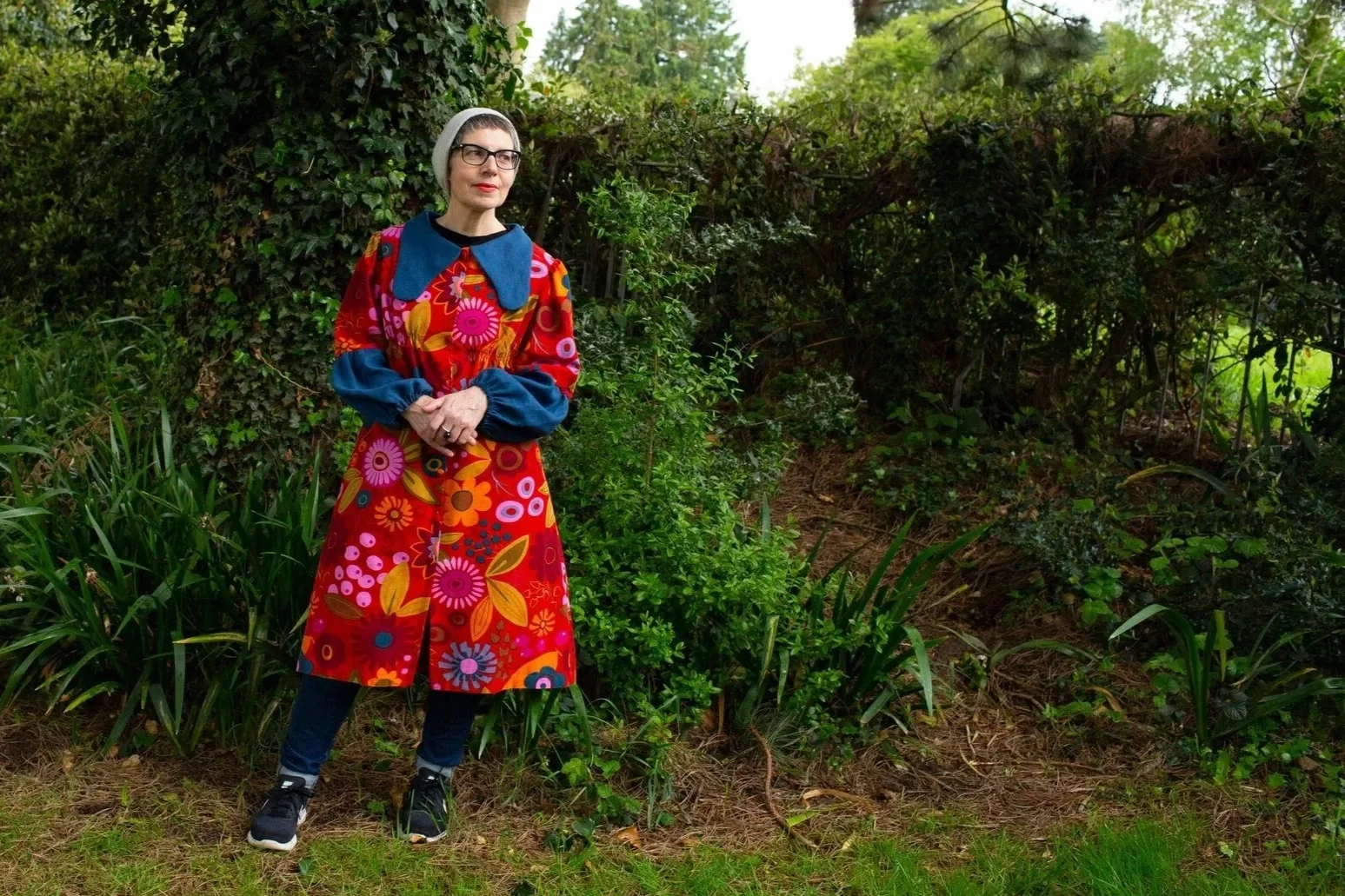 A portrait of Sonia Boué, a woman with short grey hair and glasses standing outdoors in a garden, wearing a colourful floral dress with long sleeves, a grey beanie, and black shoes.
