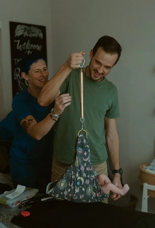 A man and woman playfully hold and lift a weighted bag with a strap, resembling a baby, with the man holding the strap and the woman touching his arm, both smiling in a room with a chalkboard sign that says 'Welcome' in the background.