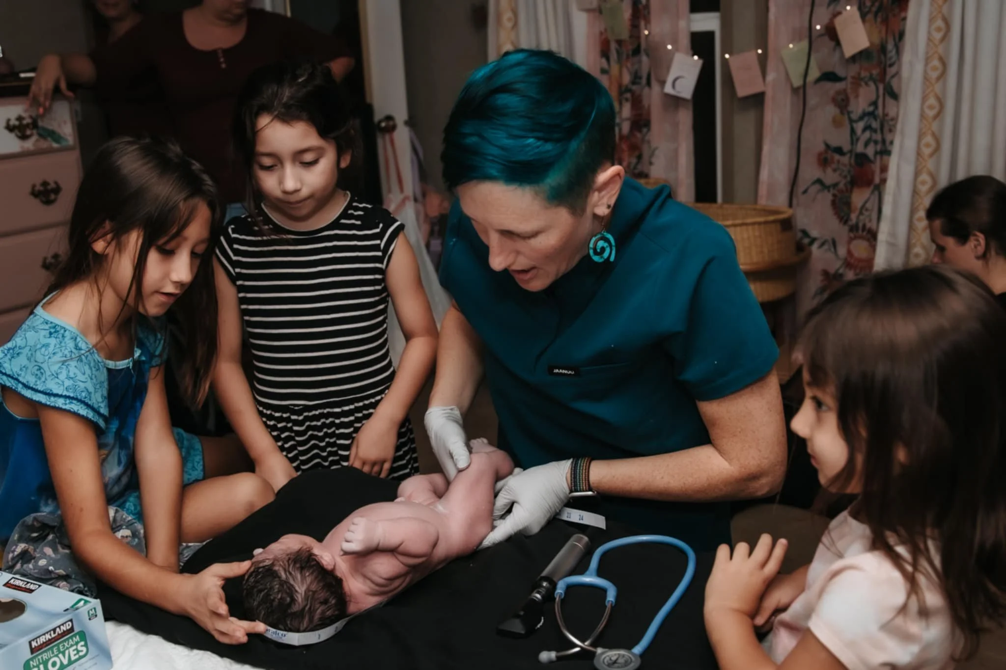 A woman with blue hair and earrings demonstrates a medical procedure on a mannequin, surrounded by children watching attentively in a room decorated with lights and cards hanging on the wall.