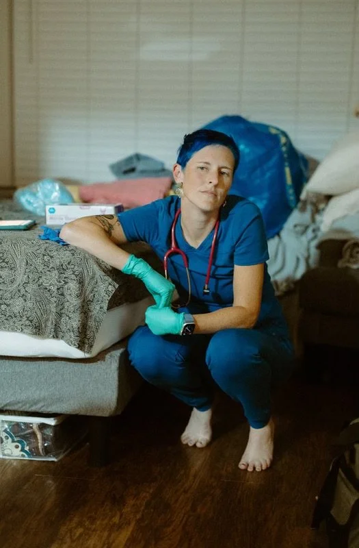 A person in blue scrubs with a stethoscope around their neck, sitting on the floor next to a bed, wearing green gloves and holding a green object.