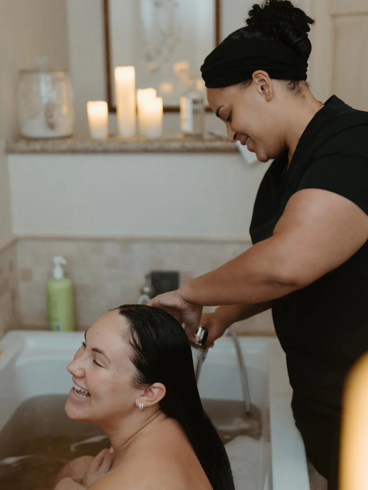 A woman is sitting in a hair washing sink with her eyes closed, smiling, while a hairstylist washes her hair.