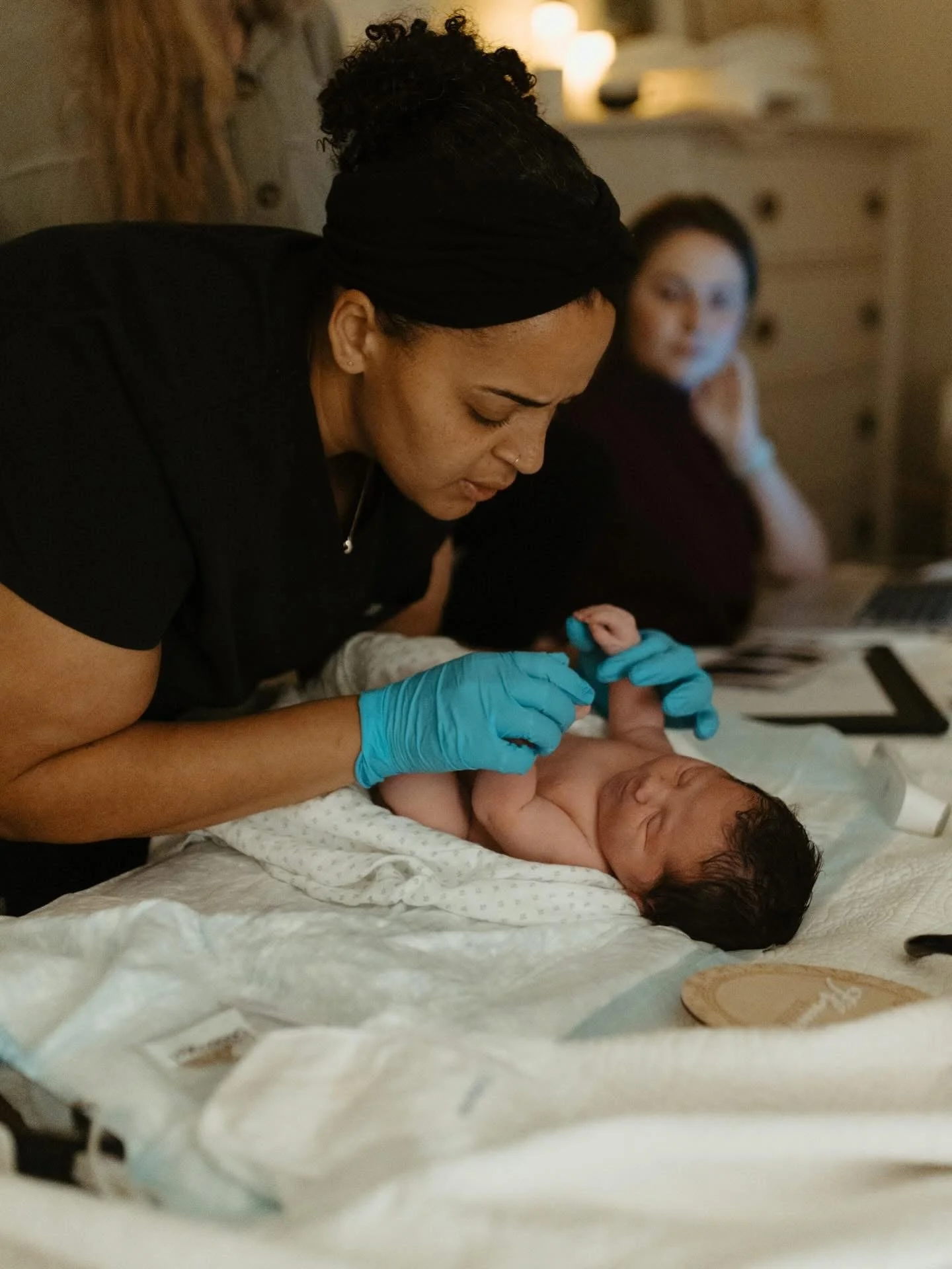 A Katy midwife, wearing blue gloves and a black headband, tending to a newborn baby lying on a bed. In the background, a woman is sitting, observing the baby.