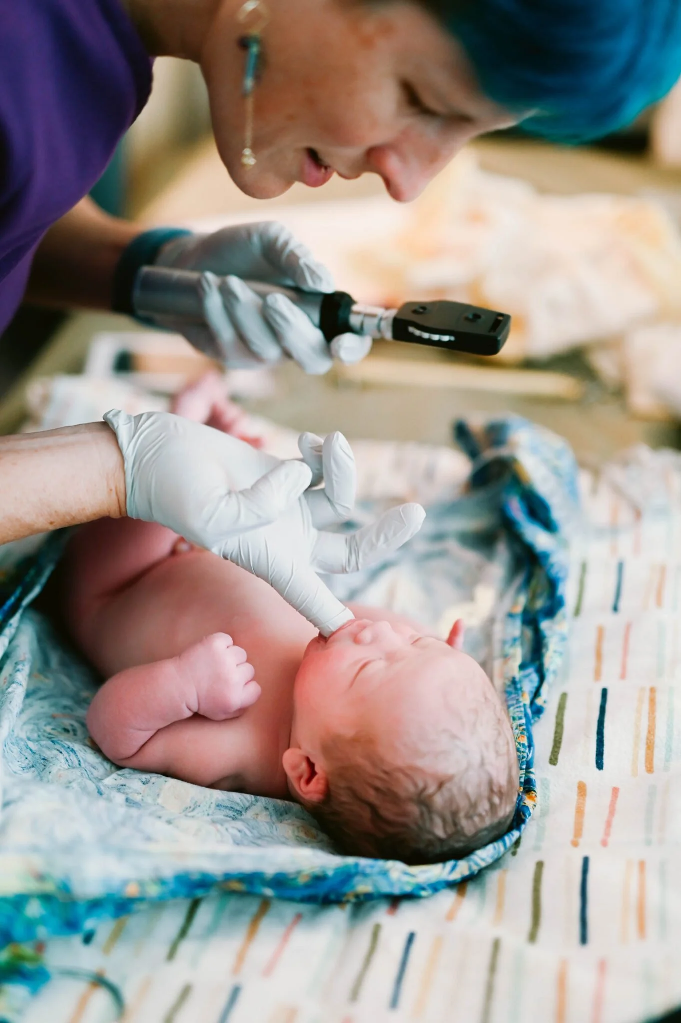 A newborn baby lying on a blanket receiving care from medical staff, including a healthcare worker using a device to check the baby's health, with the baby's umbilical cord still attached.