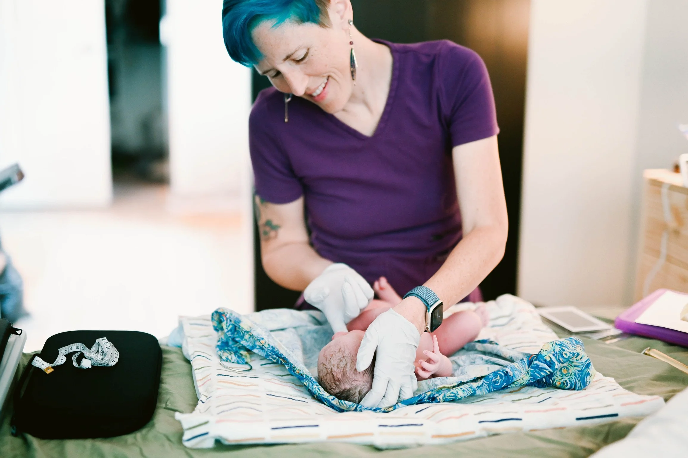 A woman with short blue hair wearing a purple shirt and white gloves is attending to a baby lying on a changing table. The woman is smiling and looking down at the baby, who is dressed in a colorful outfit. The scene appears to be in a care or medica