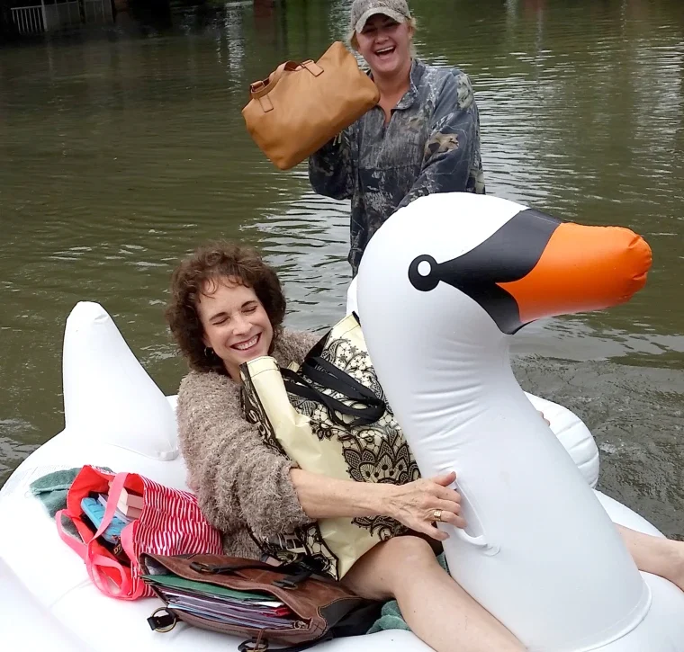Two women enjoying a boat ride on a lake. One woman is sitting in an inflatable swan with a big smile, and the other is standing outside the boat holding a brown bag, both appear happy and relaxed.