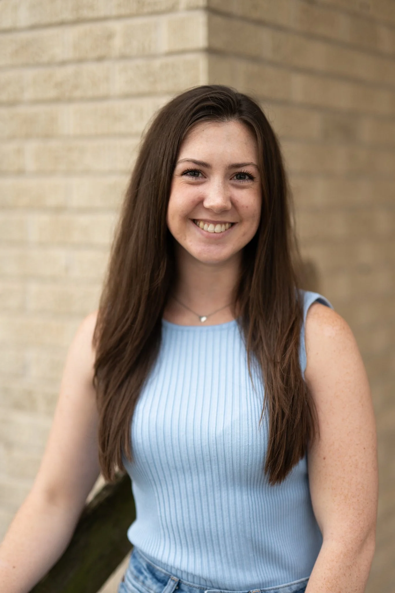 A young woman with long brown hair smiling, wearing a sleeveless light blue top with a vertical ribbed pattern, standing outdoors near a beige brick wall.